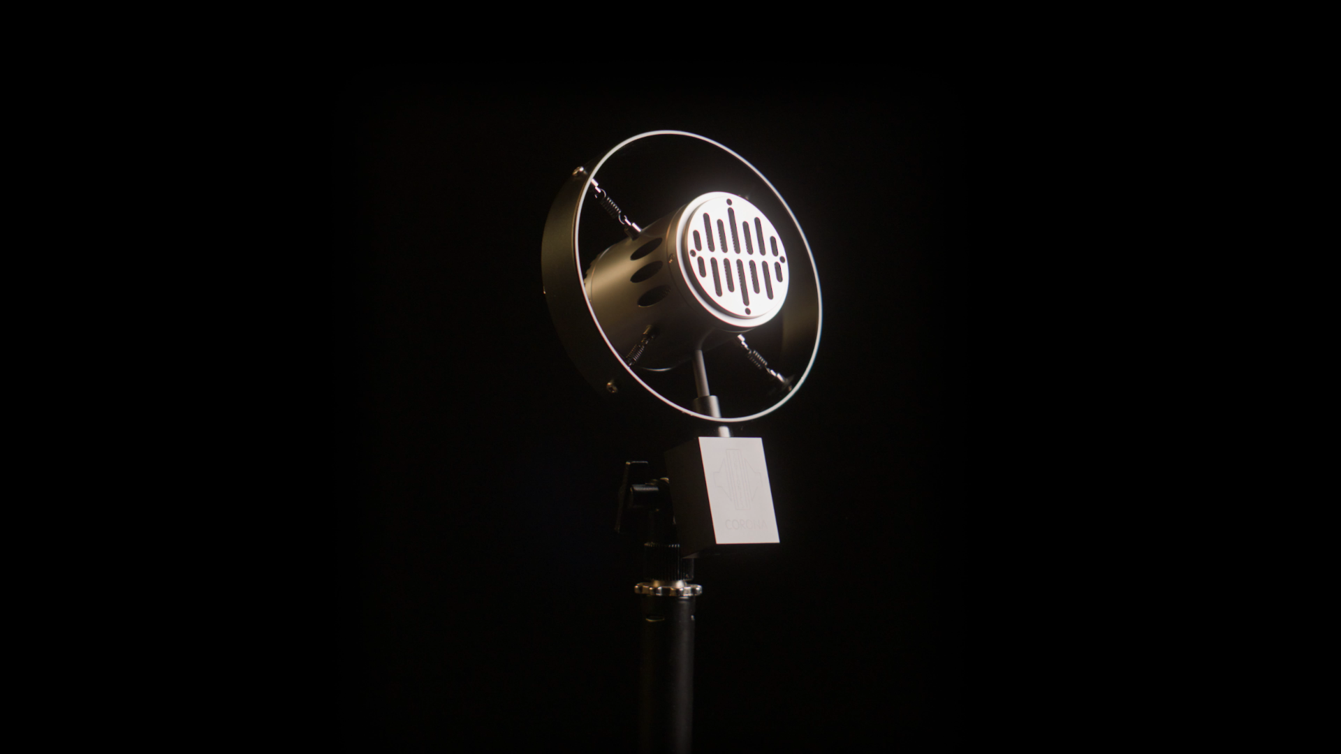 A vintage microphone with a circular grill and a metallic body, mounted on a stand against a dark background.