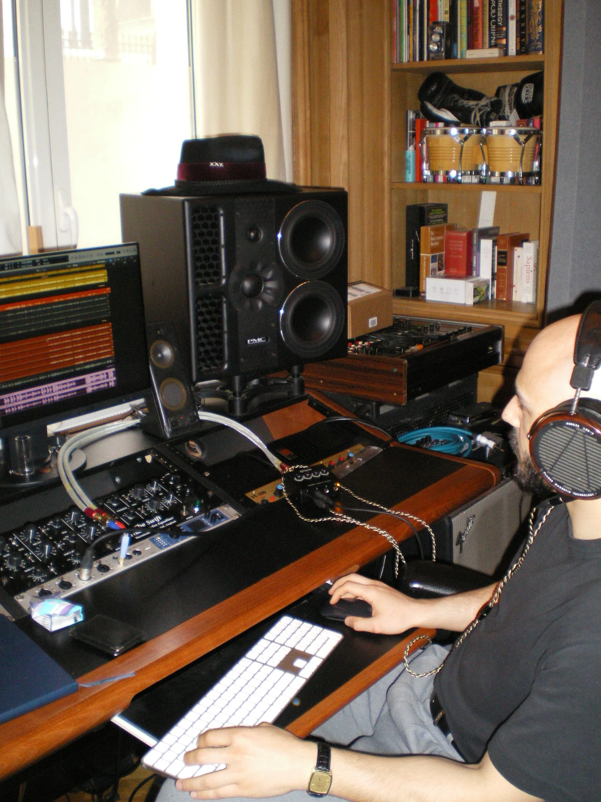 A person wearing headphones sitting at a desk with music equipment, a computer, and bookshelves in the background.
