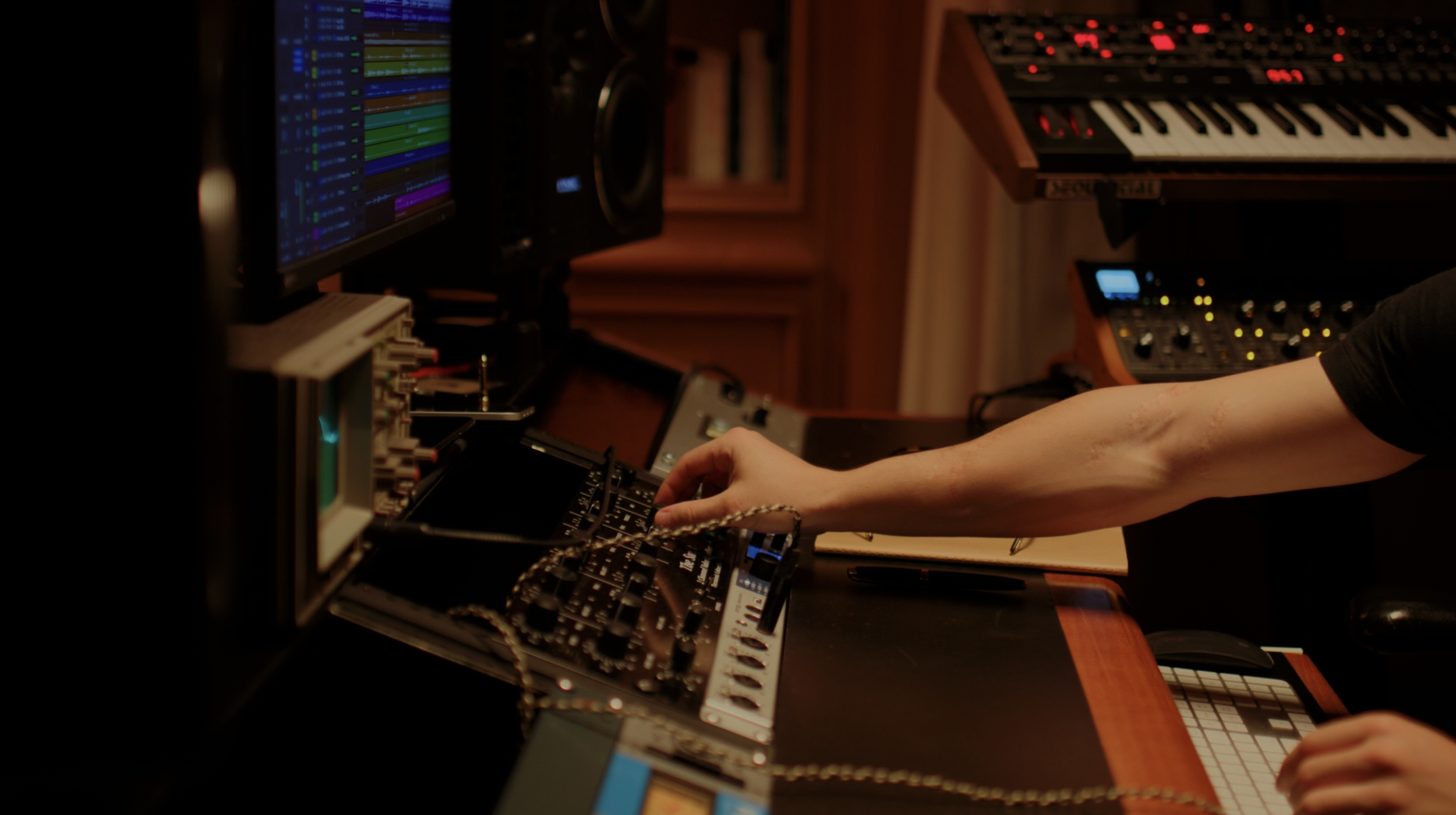 A person working in a music studio with electronic music equipment, including a computer monitor displaying digital audio software, synthesizers, and a mixing console.