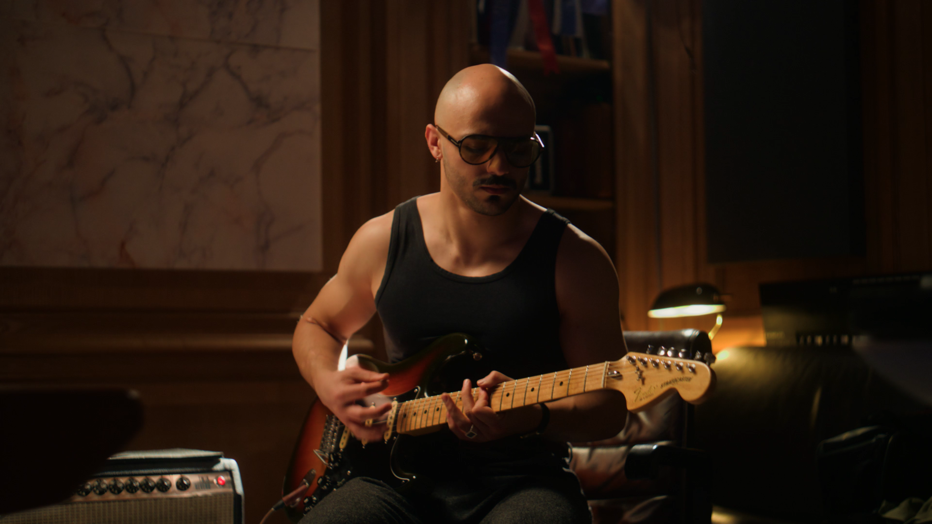 A man with a shaved head, beard, and sunglasses playing an electric guitar in a dimly lit room with wooden walls.