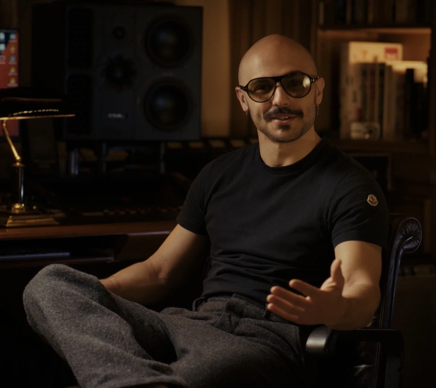 A man with a bald head, glasses, earrings, and a beard sitting in a music studio or office. He is wearing a black t-shirt and gray sweatpants, with his right arm extended. In the background, there are large speakers, a computer monitor, and wooden shelves filled with books and audio equipment.