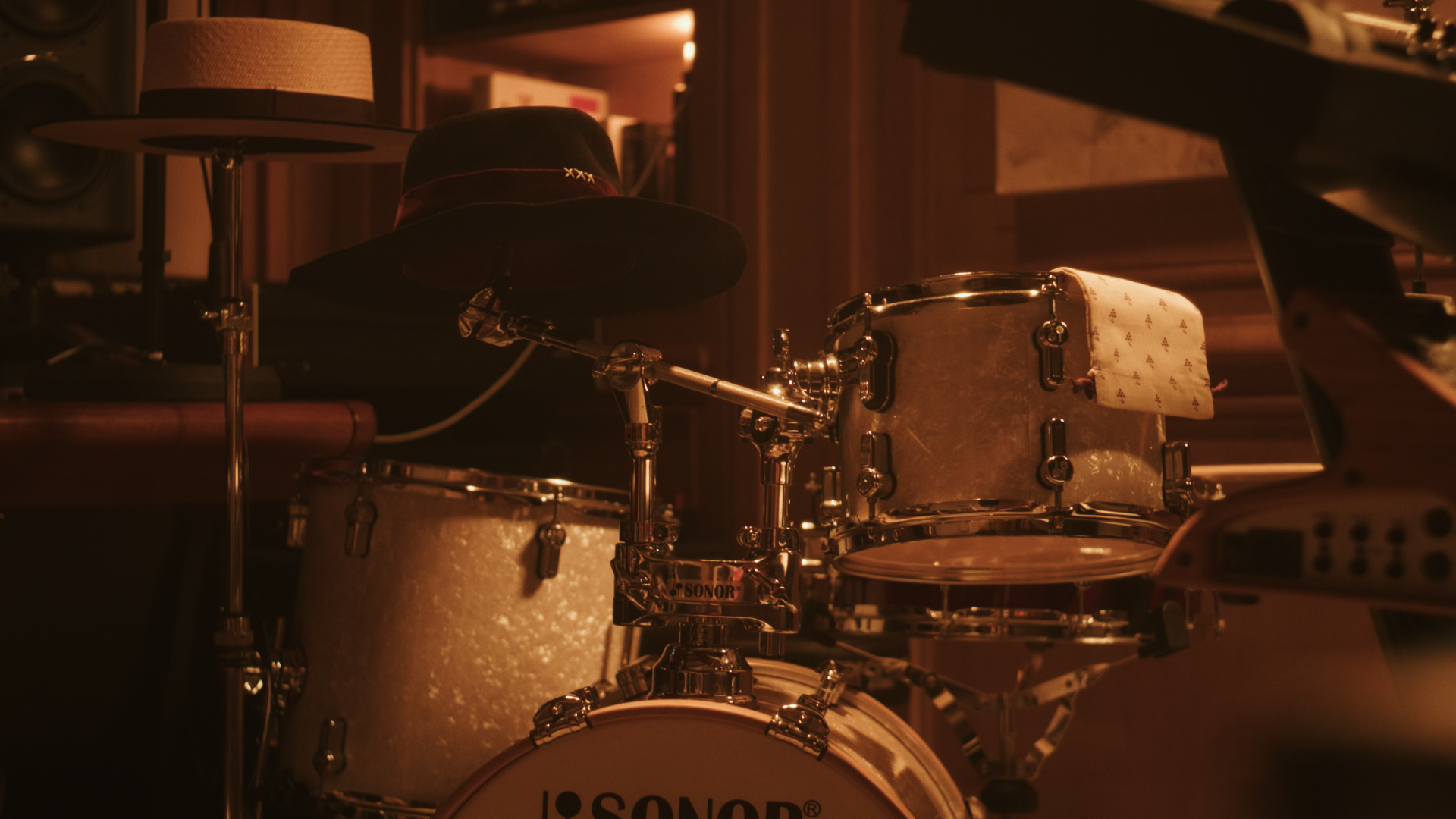A drum set with a black hat placed on top of the hi-hat, and a white cloth draped over the rack tom, in a dimly lit room.