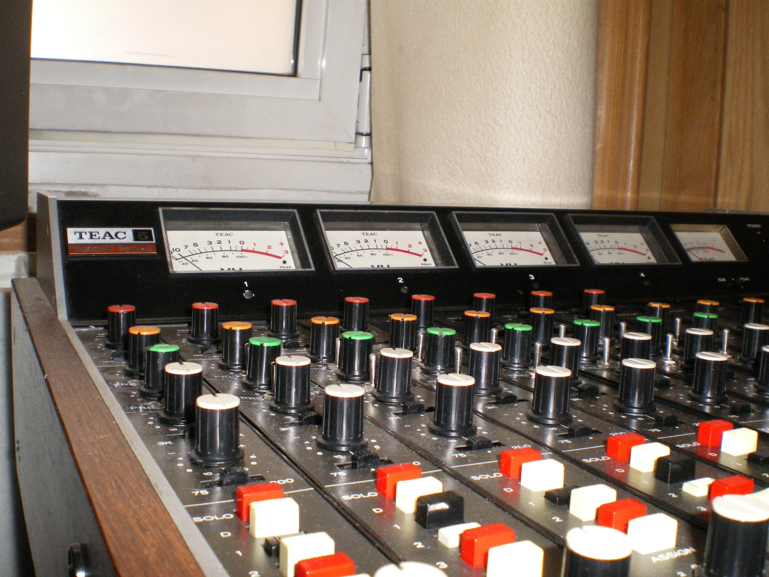 An audio mixing console with numerous colored knobs and a row of analog meters in a recording studio.