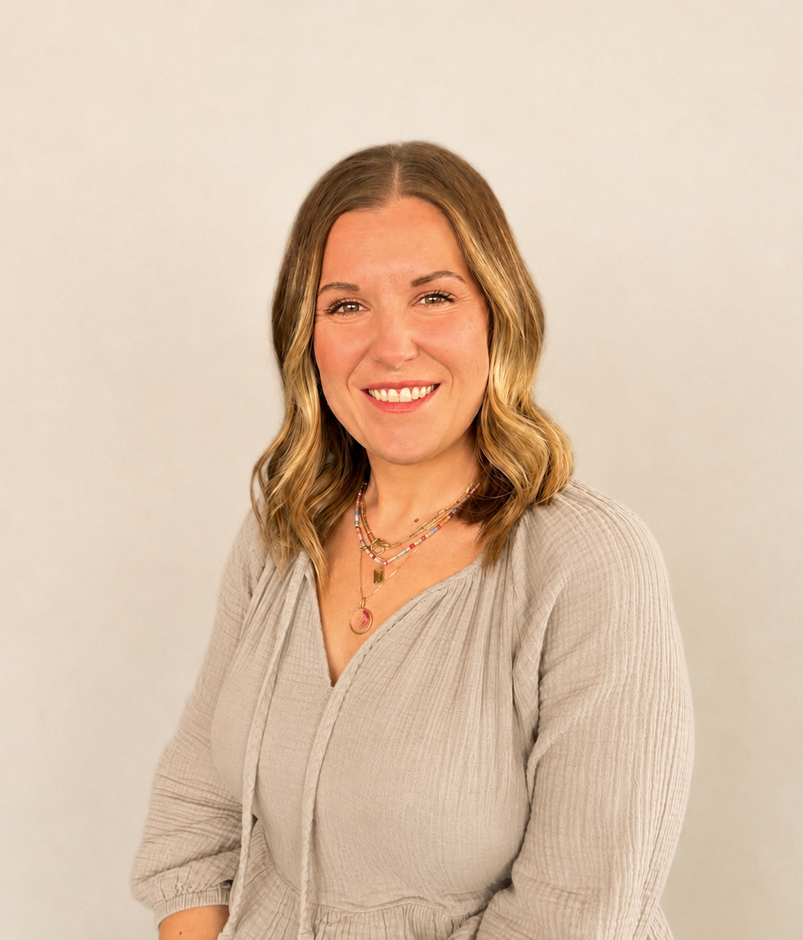 A woman with shoulder-length wavy blonde hair, wearing a beige long-sleeve blouse and layered necklaces, smiling against a plain light-colored background.