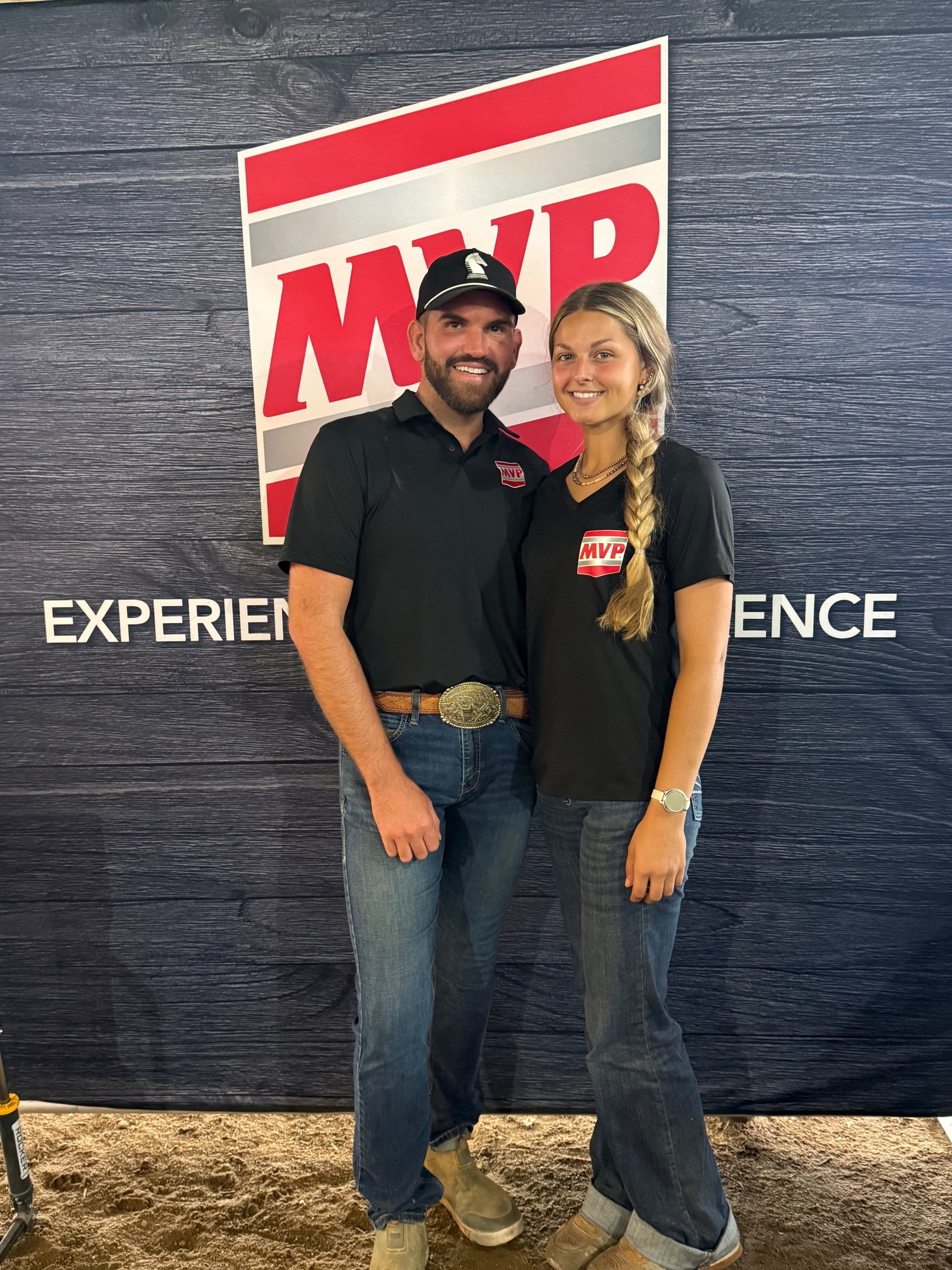 Two people, a man and a woman, standing close together and smiling at the camera in front of a black wall with a large red and white 'MVP' logo. They are both wearing black shirts with the MVP logo on the chest. The man is also wearing a black hat with a logo, jeans, and work boots. The woman has long blonde hair in a braid, is wearing a watch, layered necklaces, jeans, and boots. The ground is sandy.