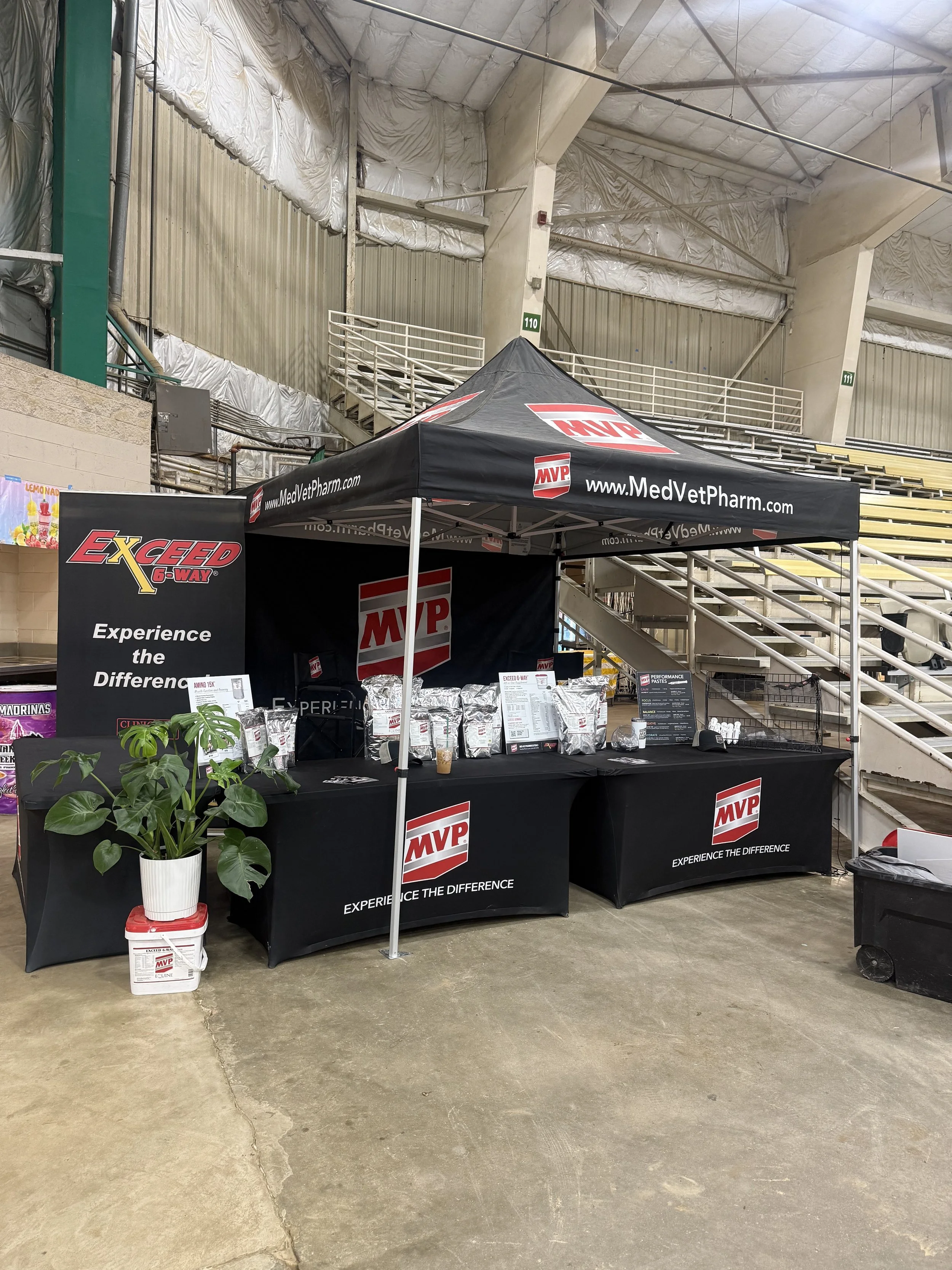 A black pavilion tent with red and white MVP logos set up inside a large, indoor sports arena. The tent has promotional banners and products displayed on tables underneath. There are steps and metallic structures in the background, with some foil insulation on the walls.