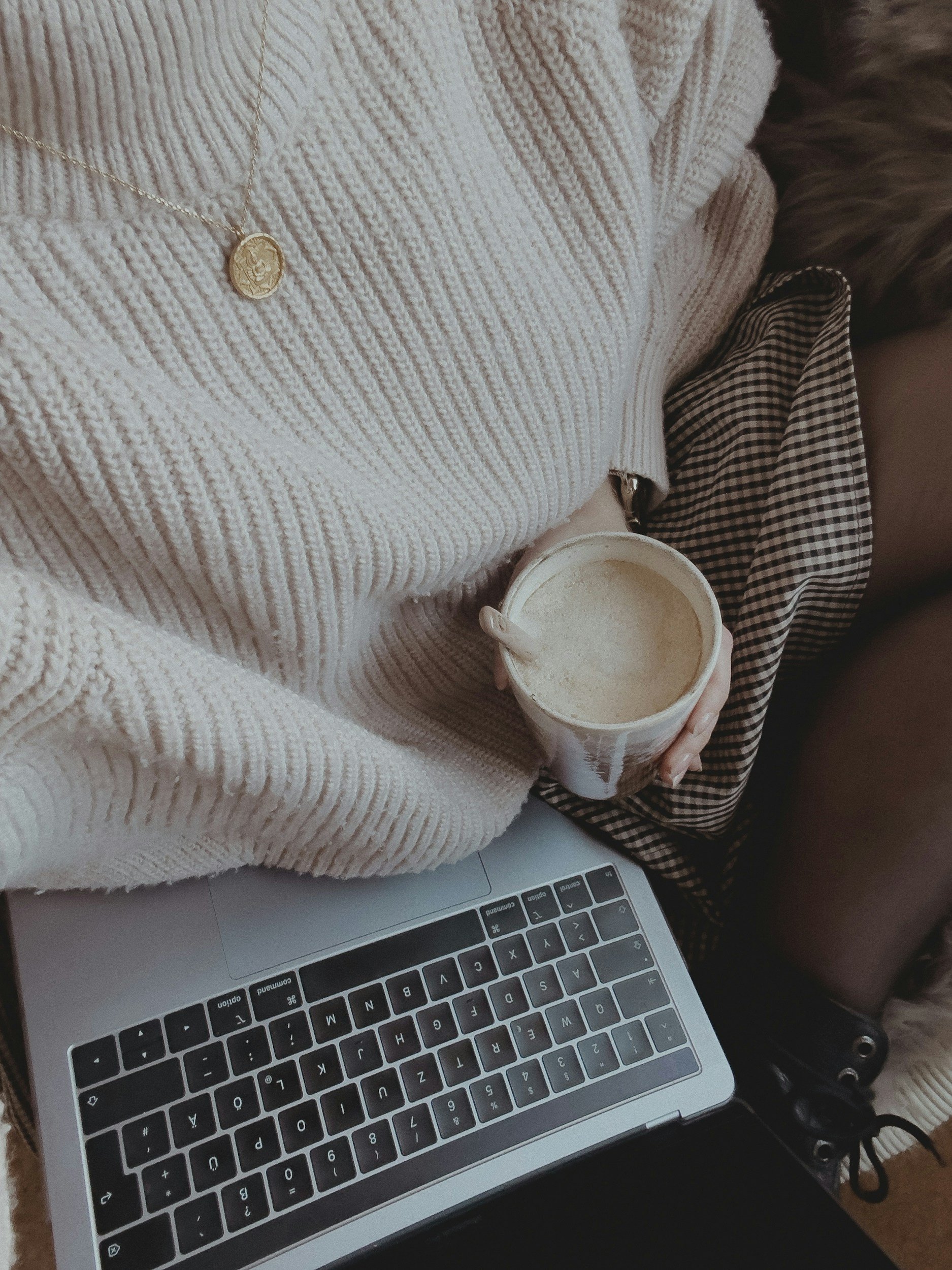 A person wearing a cream-colored knitted sweater with gold necklace holding a beige cup of coffee, with a laptop placed on their lap and a person with dark hair wearing checkered shirt beside them.