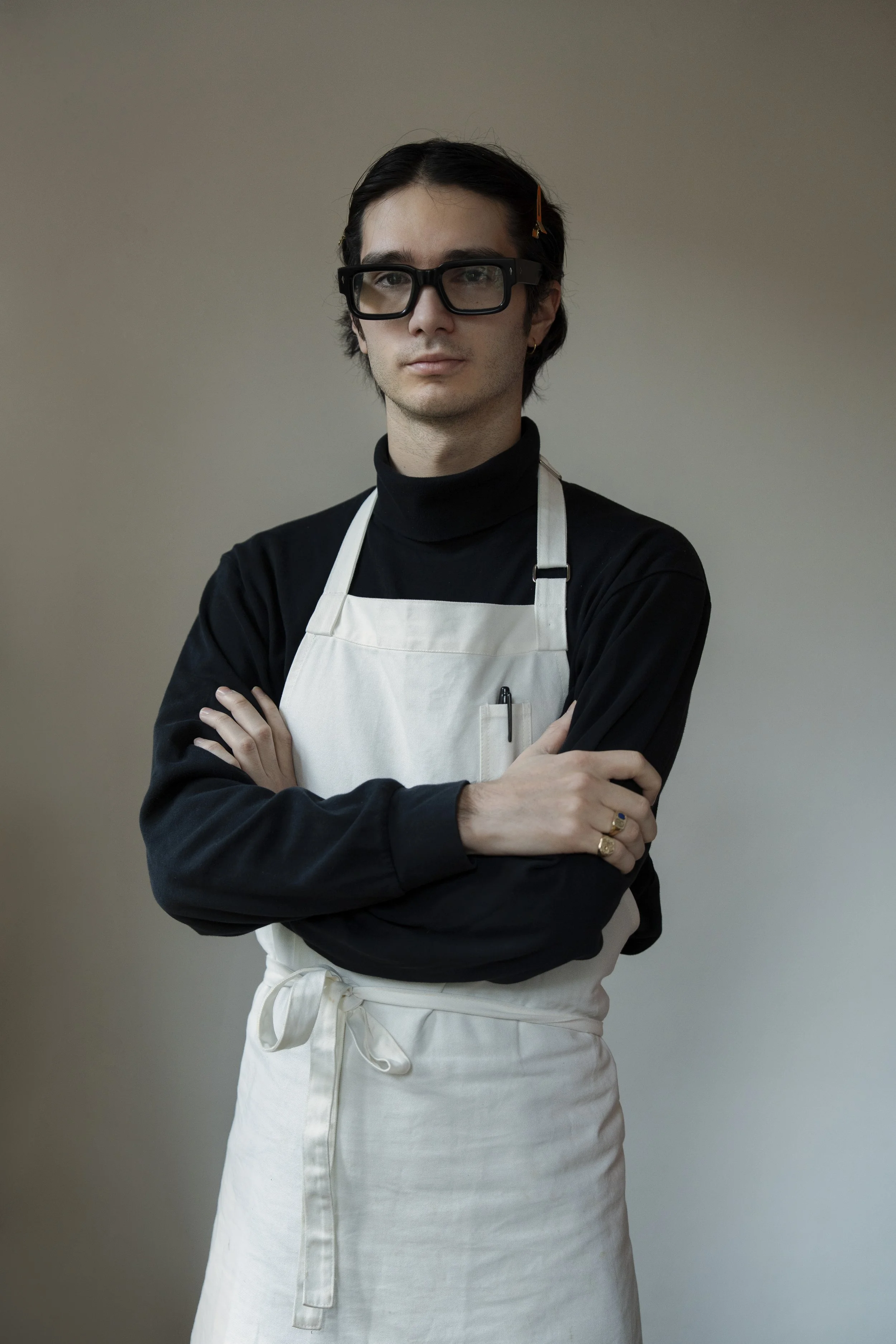 A person with dark hair, wearing black glasses, a black turtleneck, and a white apron, standing with arms crossed against a plain gray background.