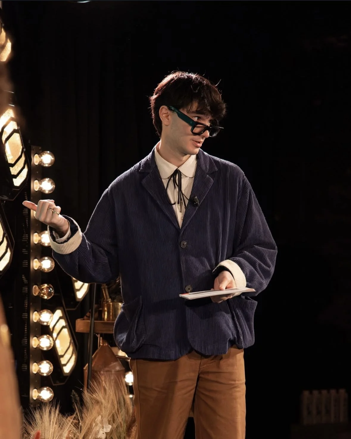 A young man with dark hair and glasses stands on a stage with black curtains, holding a notebook in his left hand and pointing with his right hand. The stage background includes decorative lights and a gold-colored object.