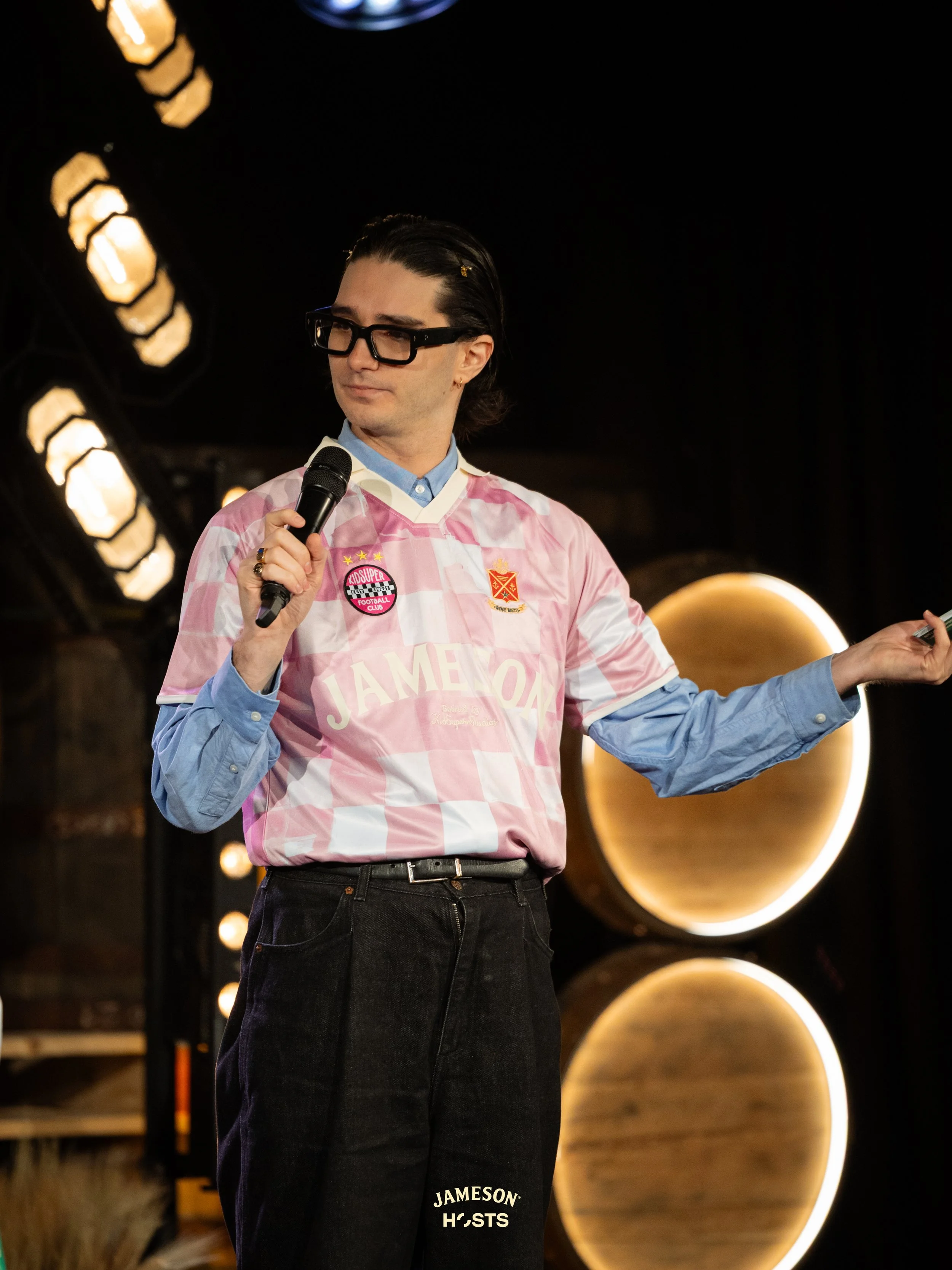 A young man with dark hair and glasses stands on a stage with black curtains, holding a notebook in his left hand and pointing with his right hand. The stage background includes decorative lights and a gold-colored object.