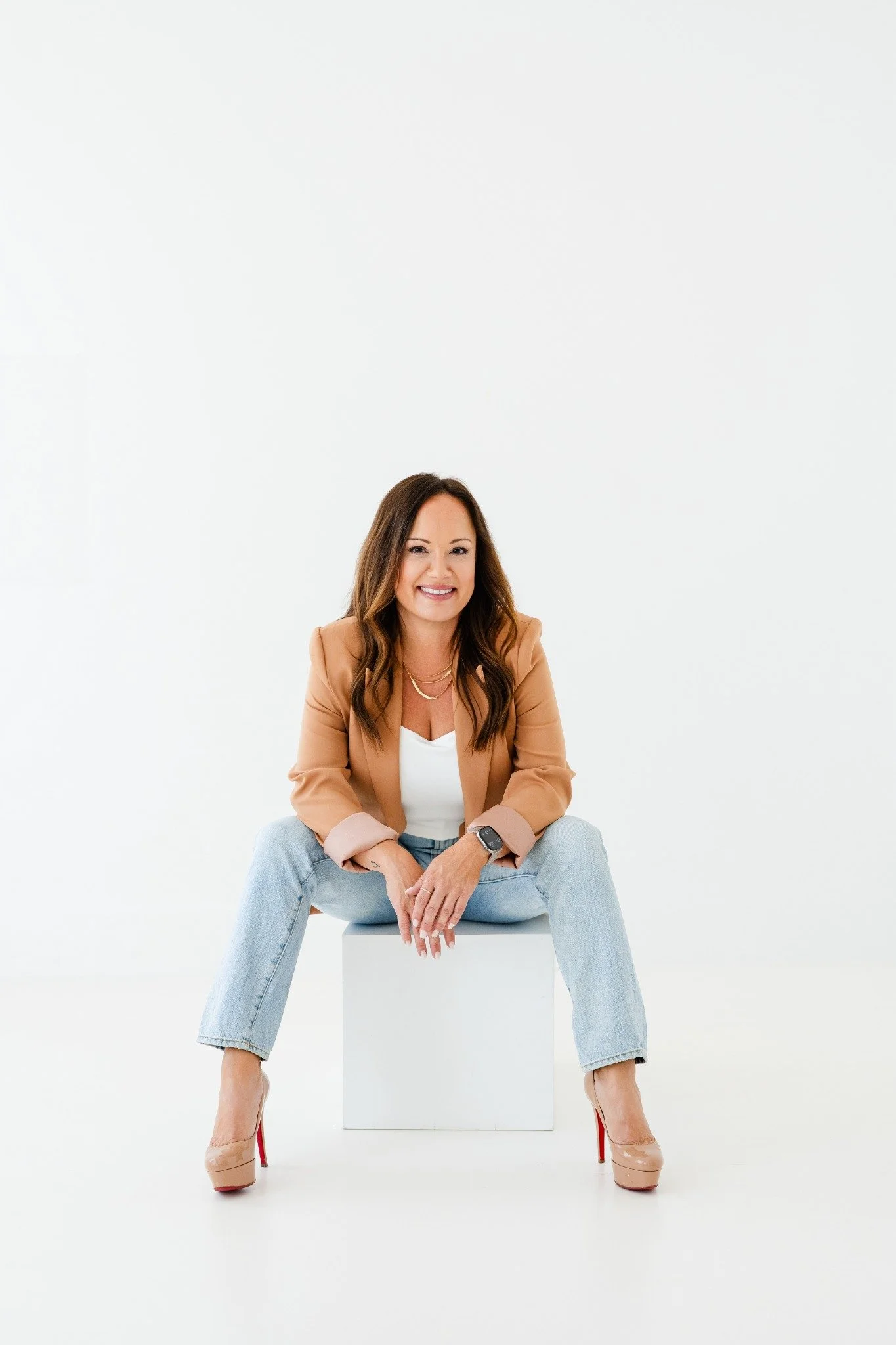 A woman with brown hair sitting on a white cube, smiling at the camera, dressed in a tan blazer, white top, light blue jeans, and red high heels, against a white background.