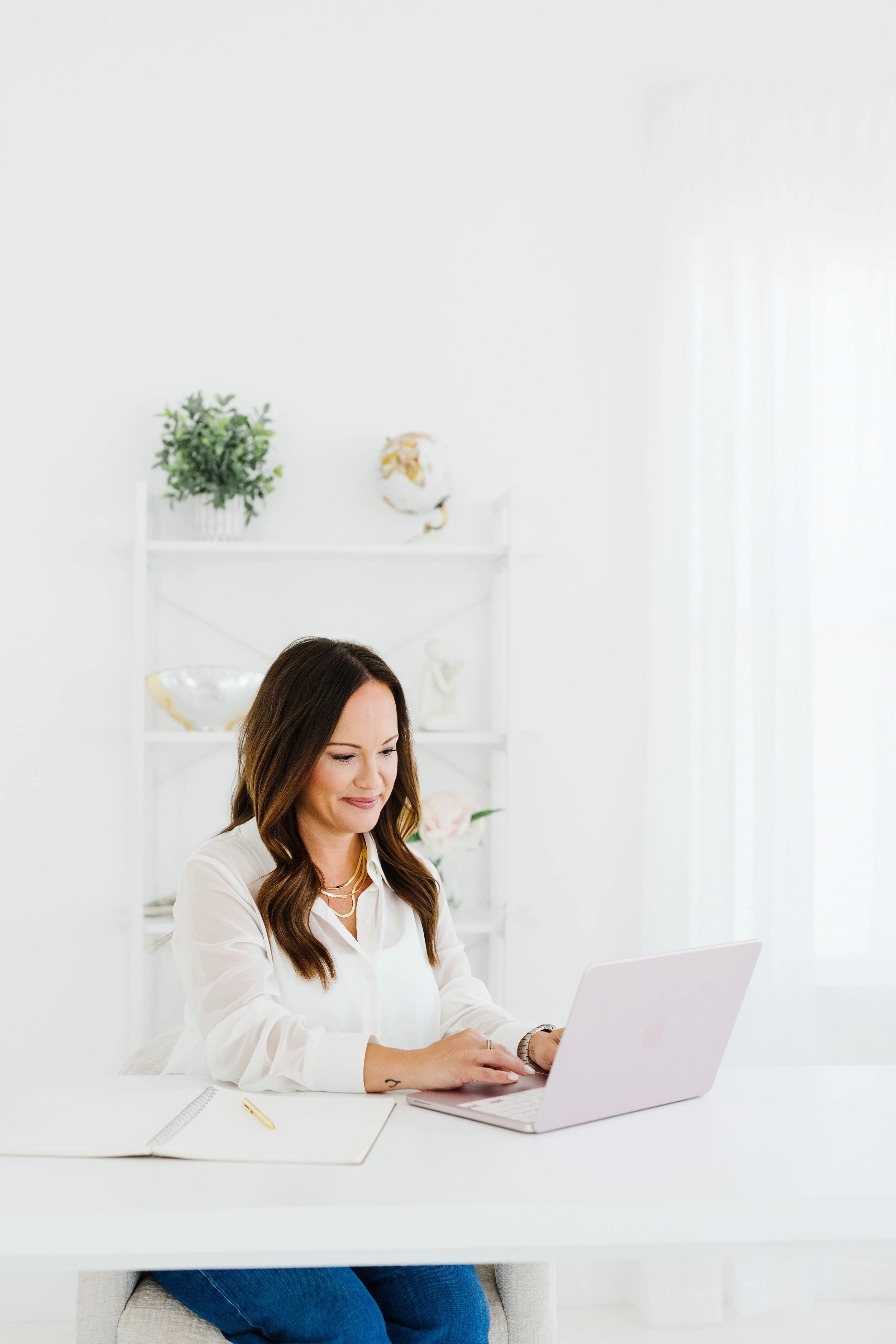 Woman with long brown hair, wearing a white blouse and gold necklace, sitting at a white desk working on a pink laptop in a bright, minimalist room with white walls and shelves decorated with plants and decorative objects.