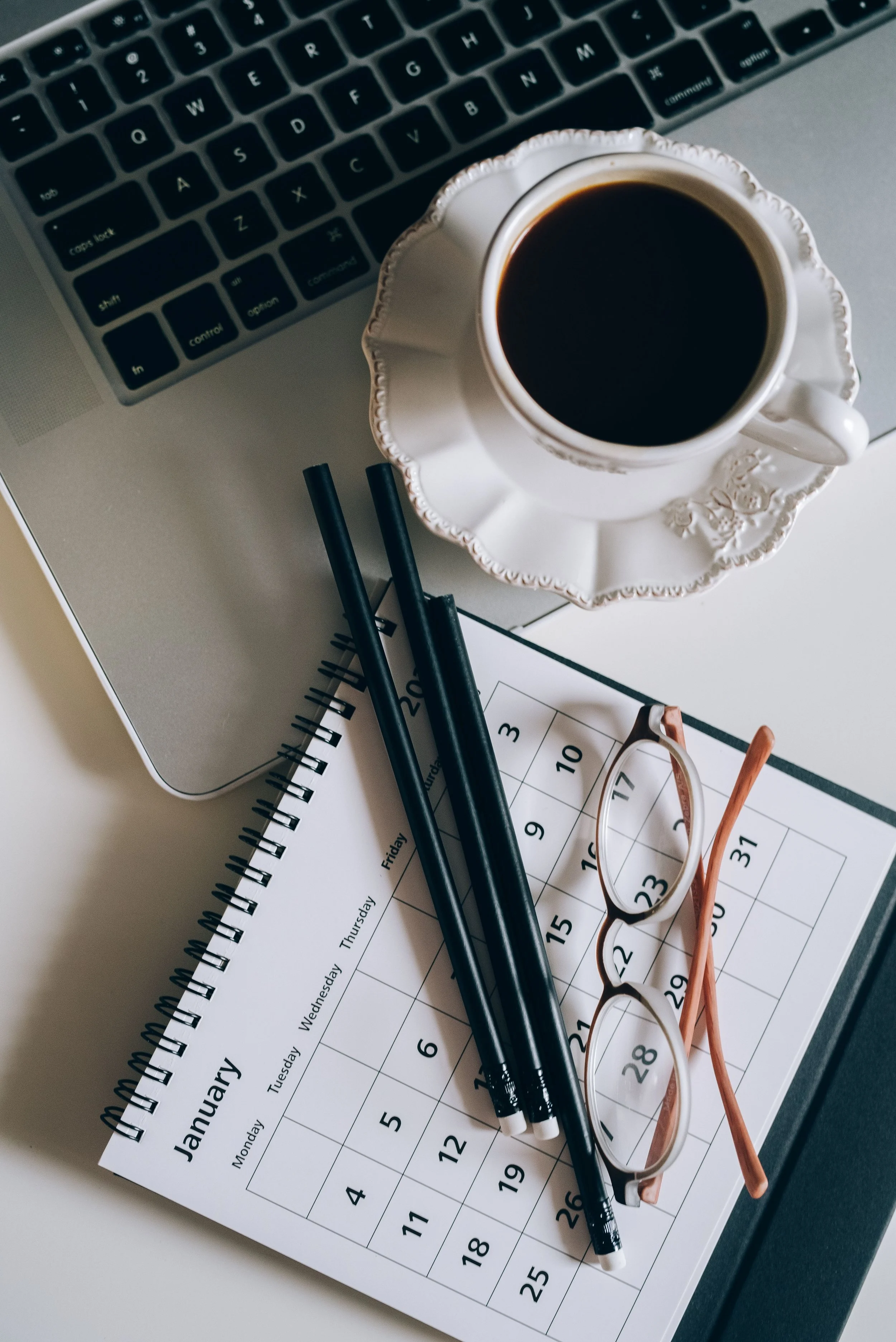 A workspace with a silver laptop keyboard, a white ornate coffee cup with black coffee, two black pens, a pair of eyeglasses, a small spiral-bound notebook, and a calendar showing days of the week and dates.