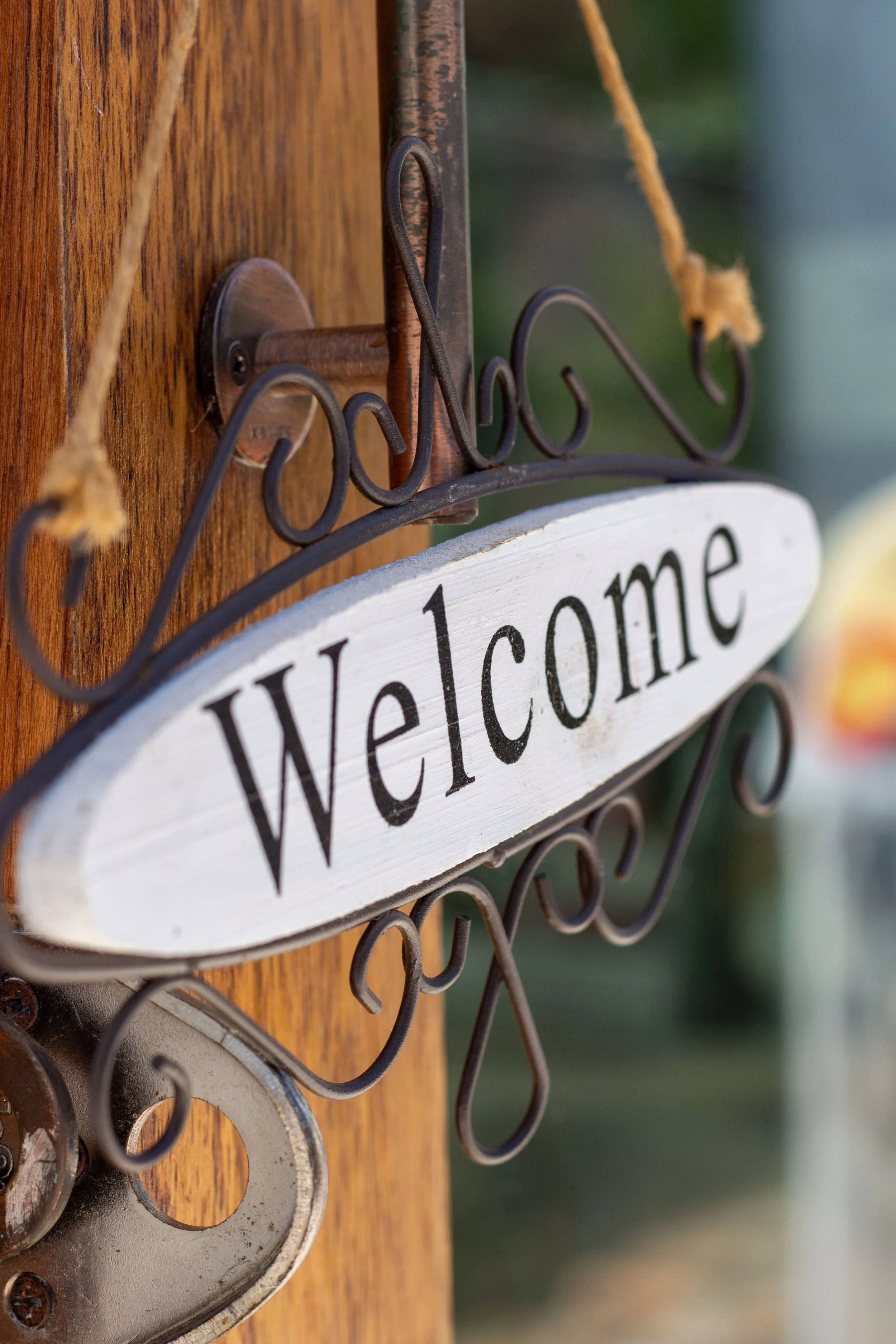Close-up photo of a wooden sign with the word 'Welcome' in black letters, mounted on a wooden post with metal hardware, and a blurred background.