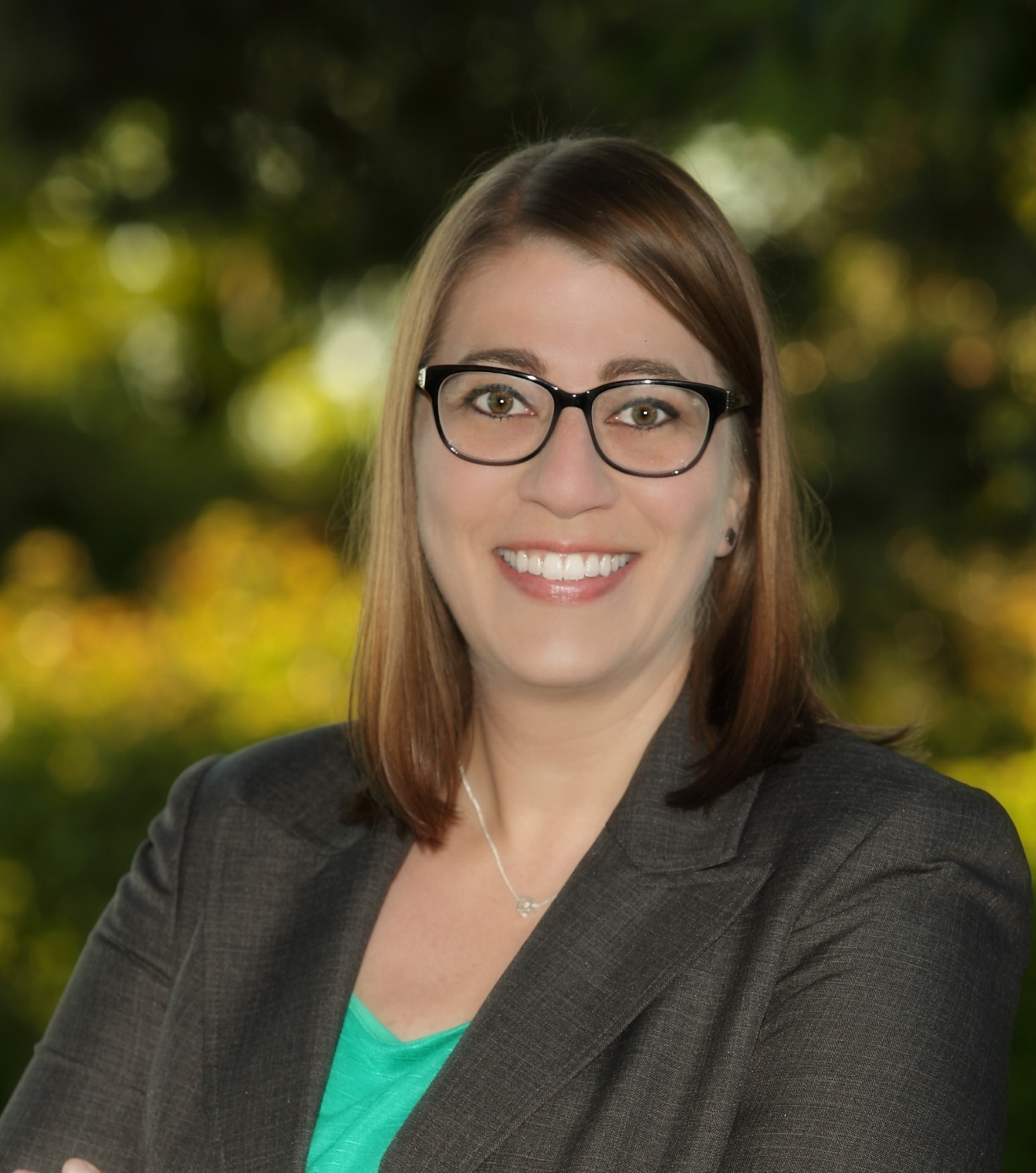 Close-up of a smiling woman with brown hair, glasses, and a black blazer, outdoors with blurred green foliage in the background.