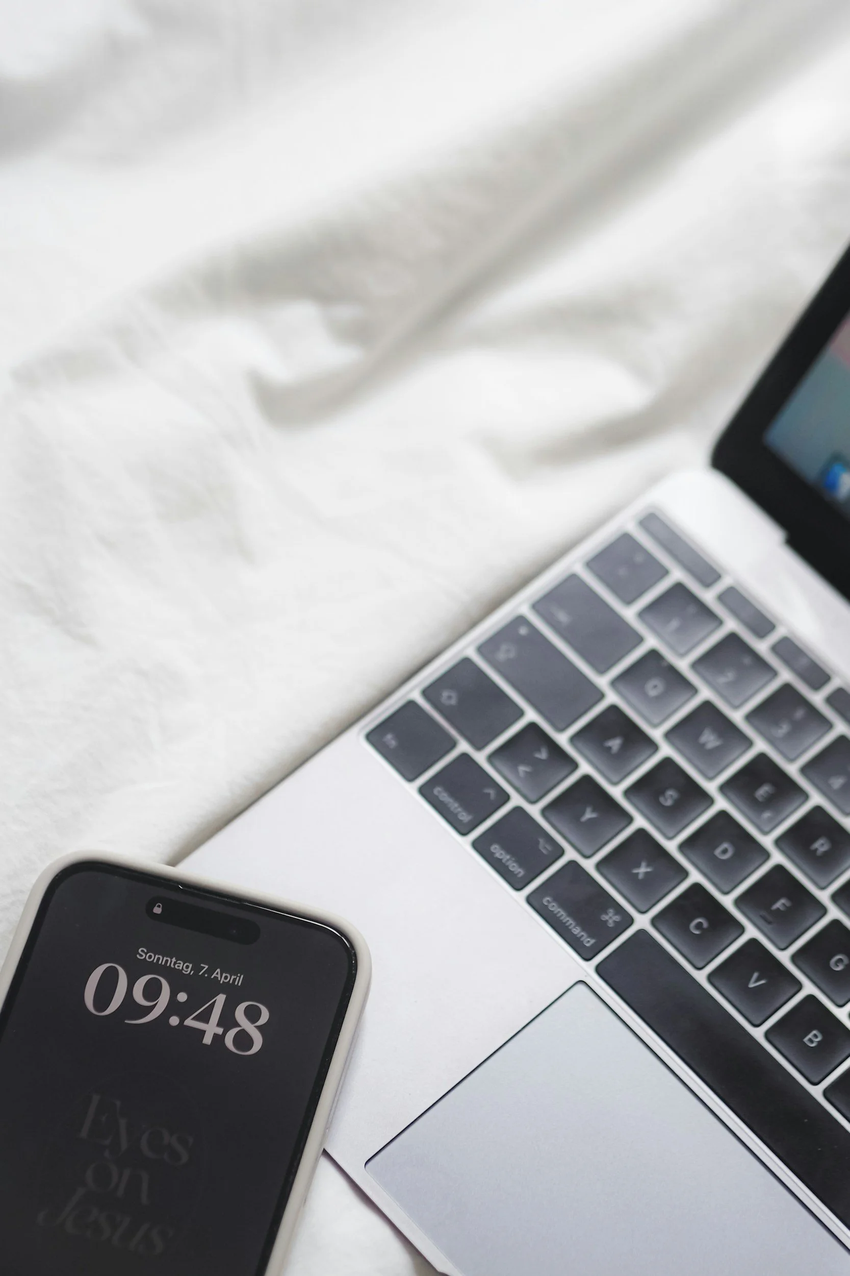A smartphone showing the date and time, a partial laptop keyboard, and a corner of a tablet or another device on a white surface.