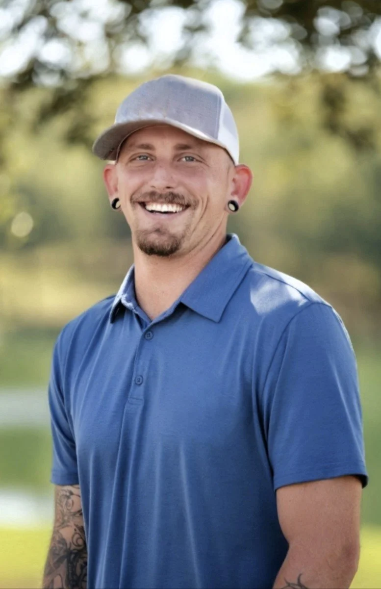 Smiling man with tattoos, earrings, and a gray cap standing outdoors in a blue polo shirt, blurred green and yellow background.