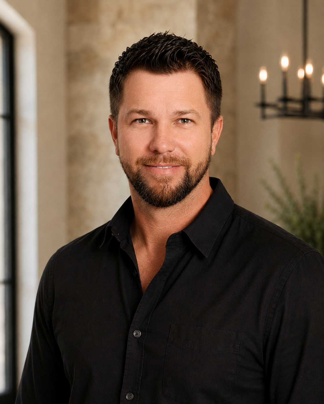 A man with short dark hair and a beard, wearing a black collared shirt, smiling indoors near a window and a chandelier with candles.