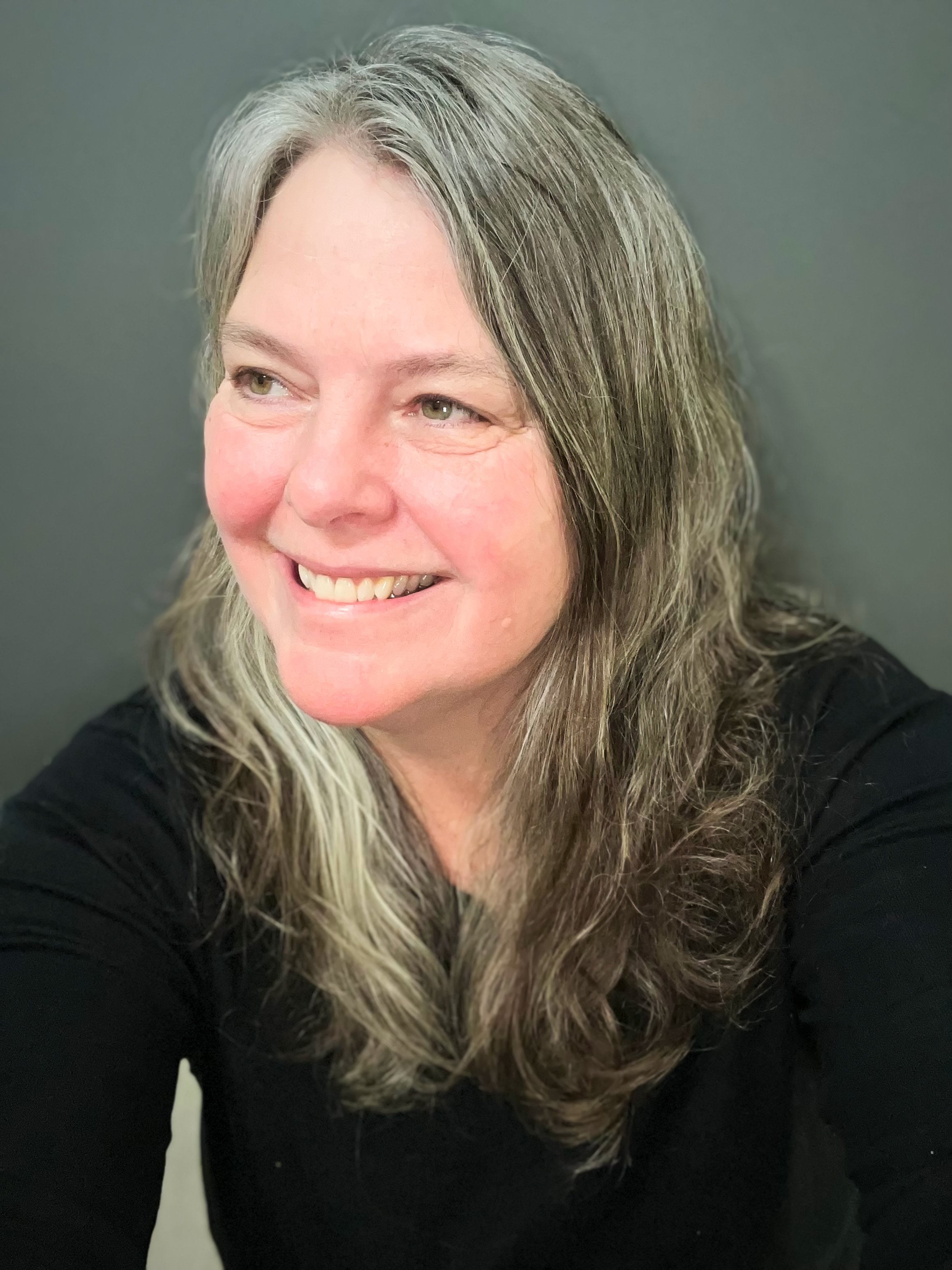 A woman with long, wavy, gray-blonde hair smiling, wearing a black top against a plain gray background.