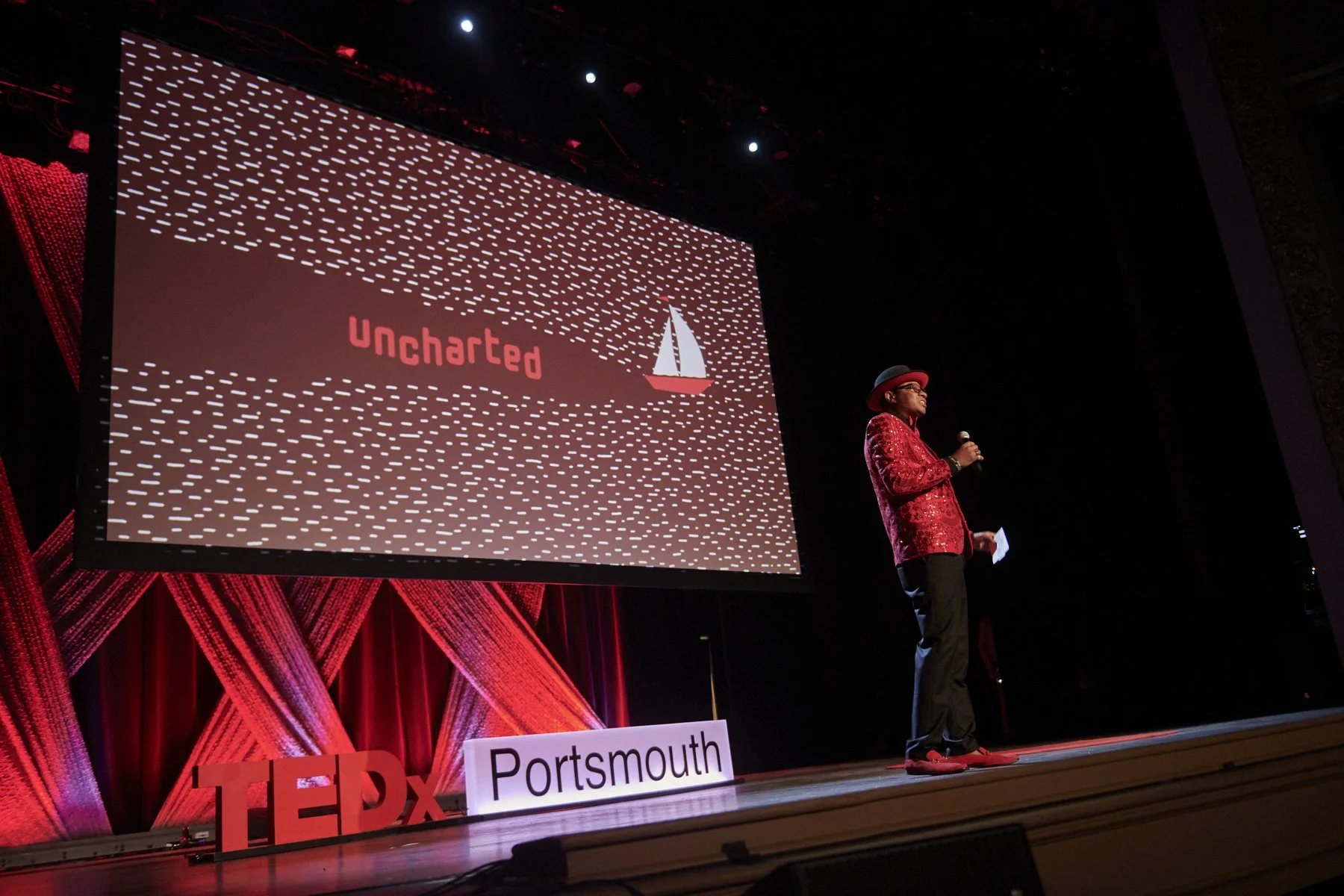 A person speaking at a TEDxPortsmouth event, standing on stage with a large screen behind displaying the word 'Uncharted' and a sailboat icon, with red and black decor.