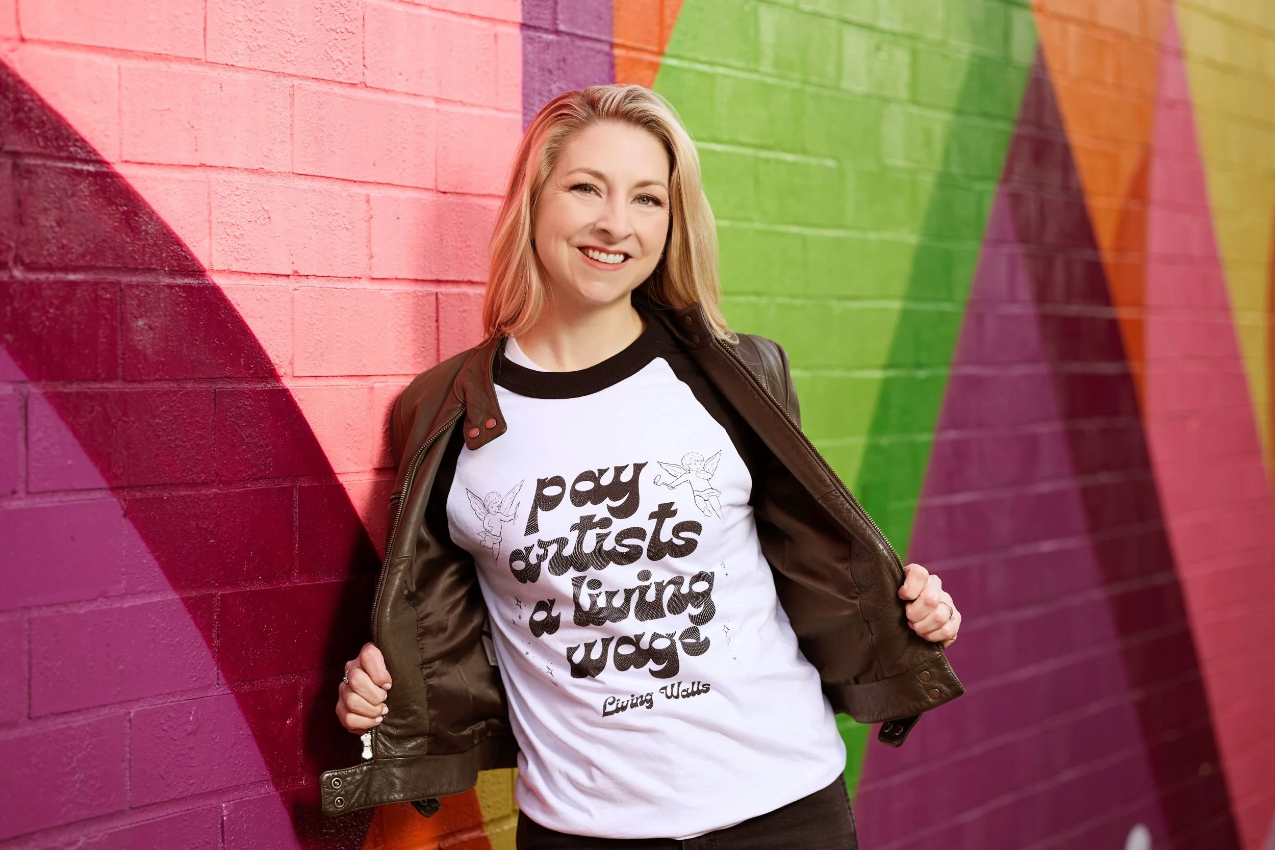 Stacy Handler in a "Pay artists a living wage" T-shirt in front of a colorful painted wall.