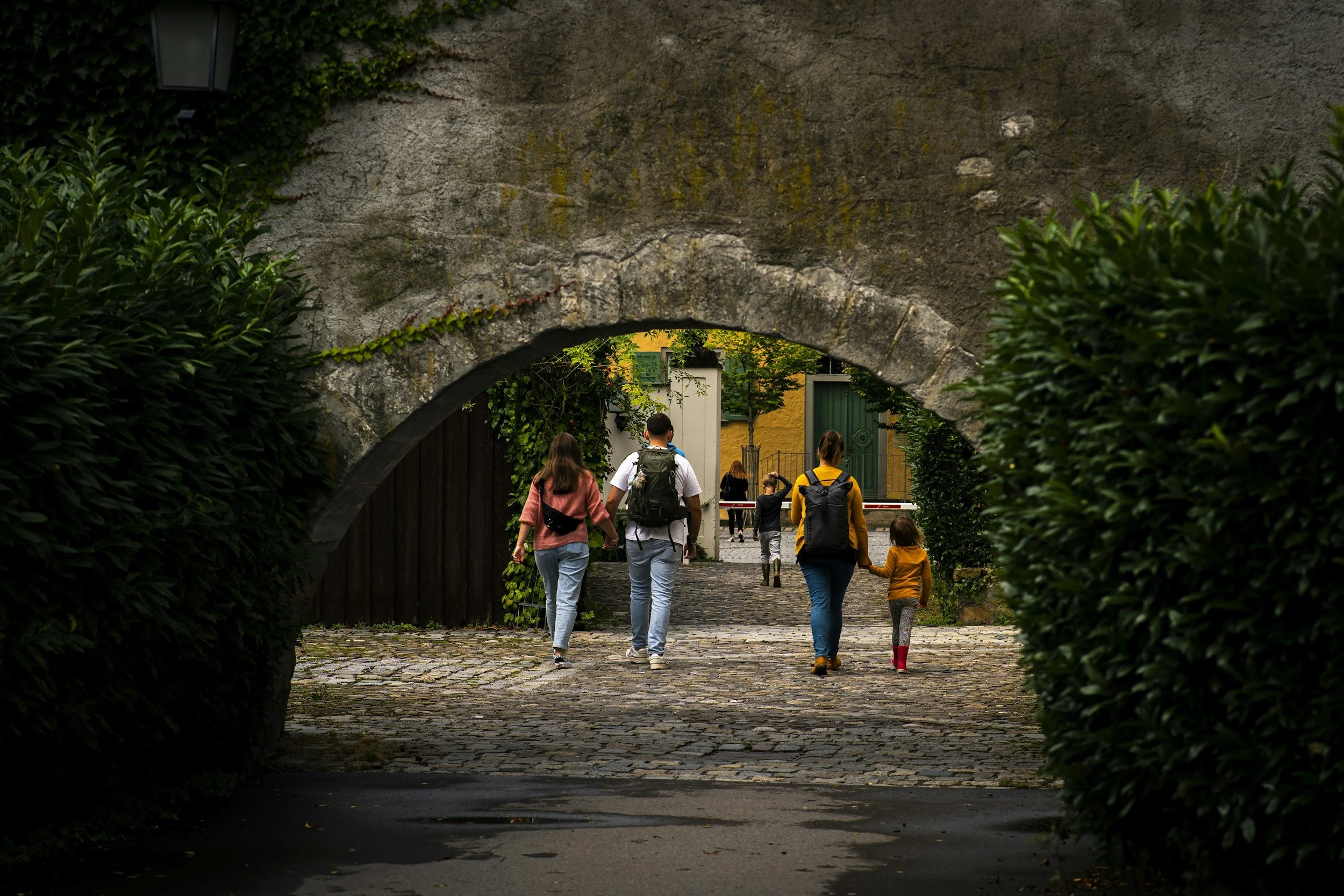 People walking through an old stone archway into a cobblestone courtyard with greenery and buildings in the background.