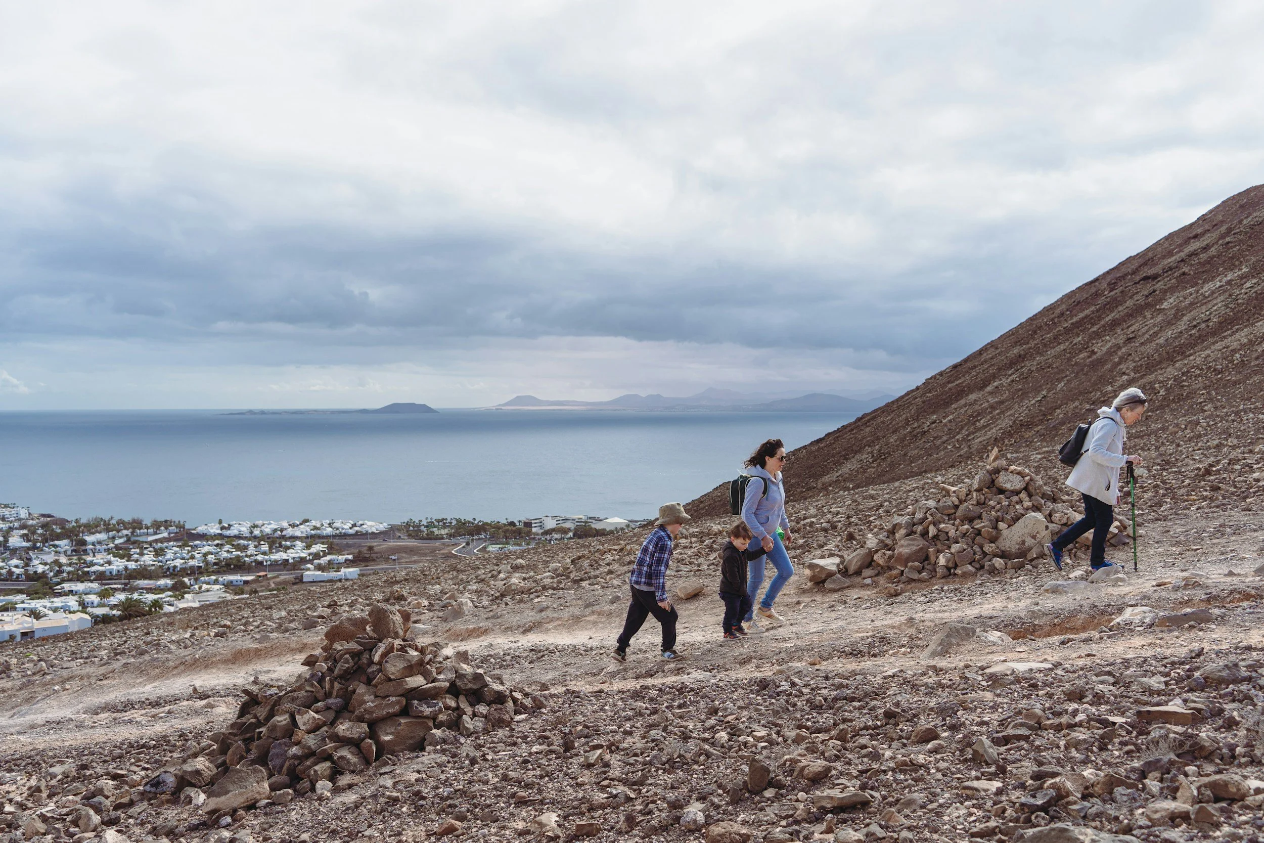 Five people hiking up a rocky path on a hillside with a coastal town below and a body of water with islands in the distance under overcast skies.