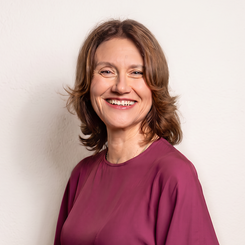 A woman with shoulder-length light brown hair, smiling and wearing a burgundy blouse, standing against a plain white wall.