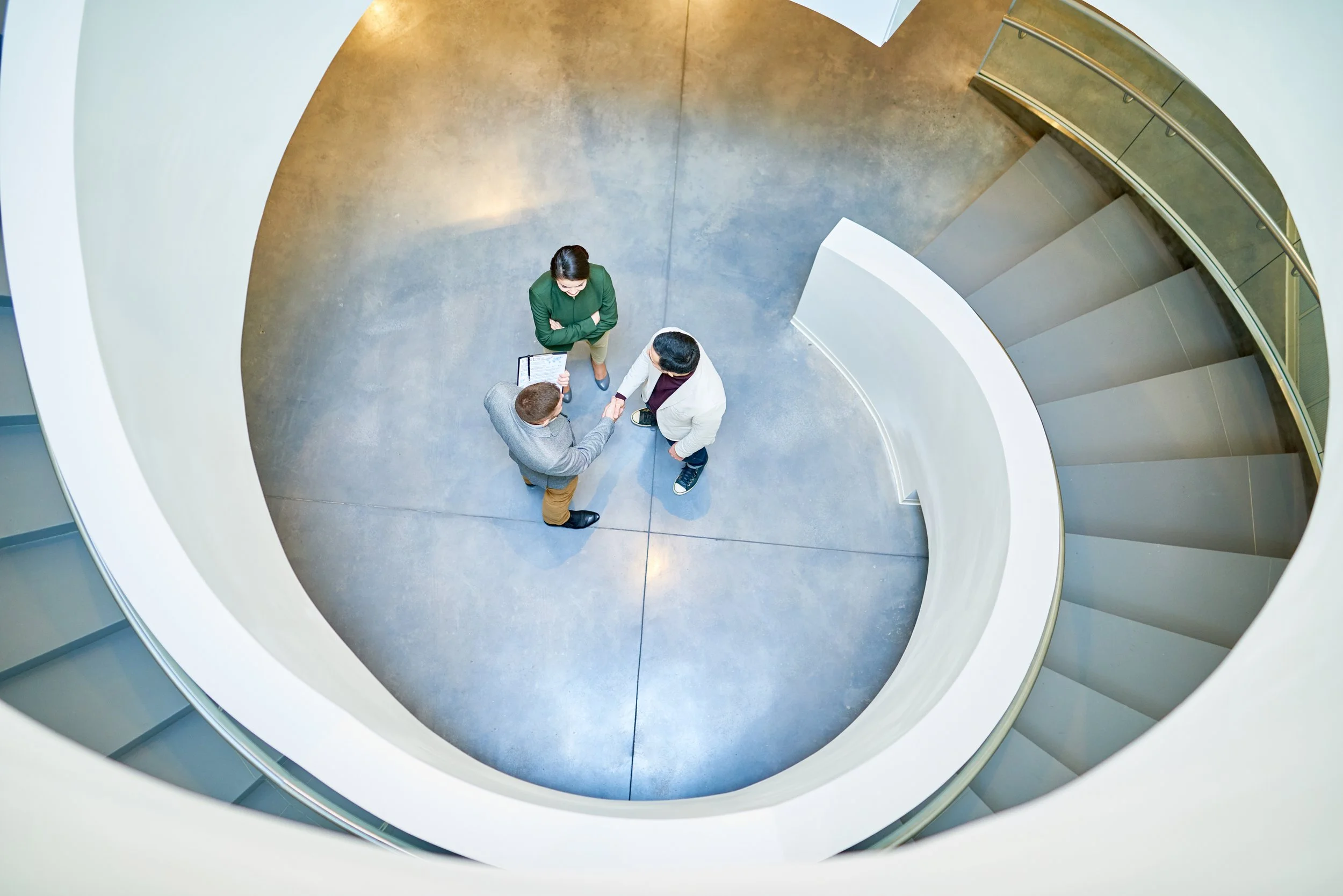 Top-down view of three people standing on a circular balcony, shaking hands, with a blank clipboard in hand, overlooking an open spiral staircase.