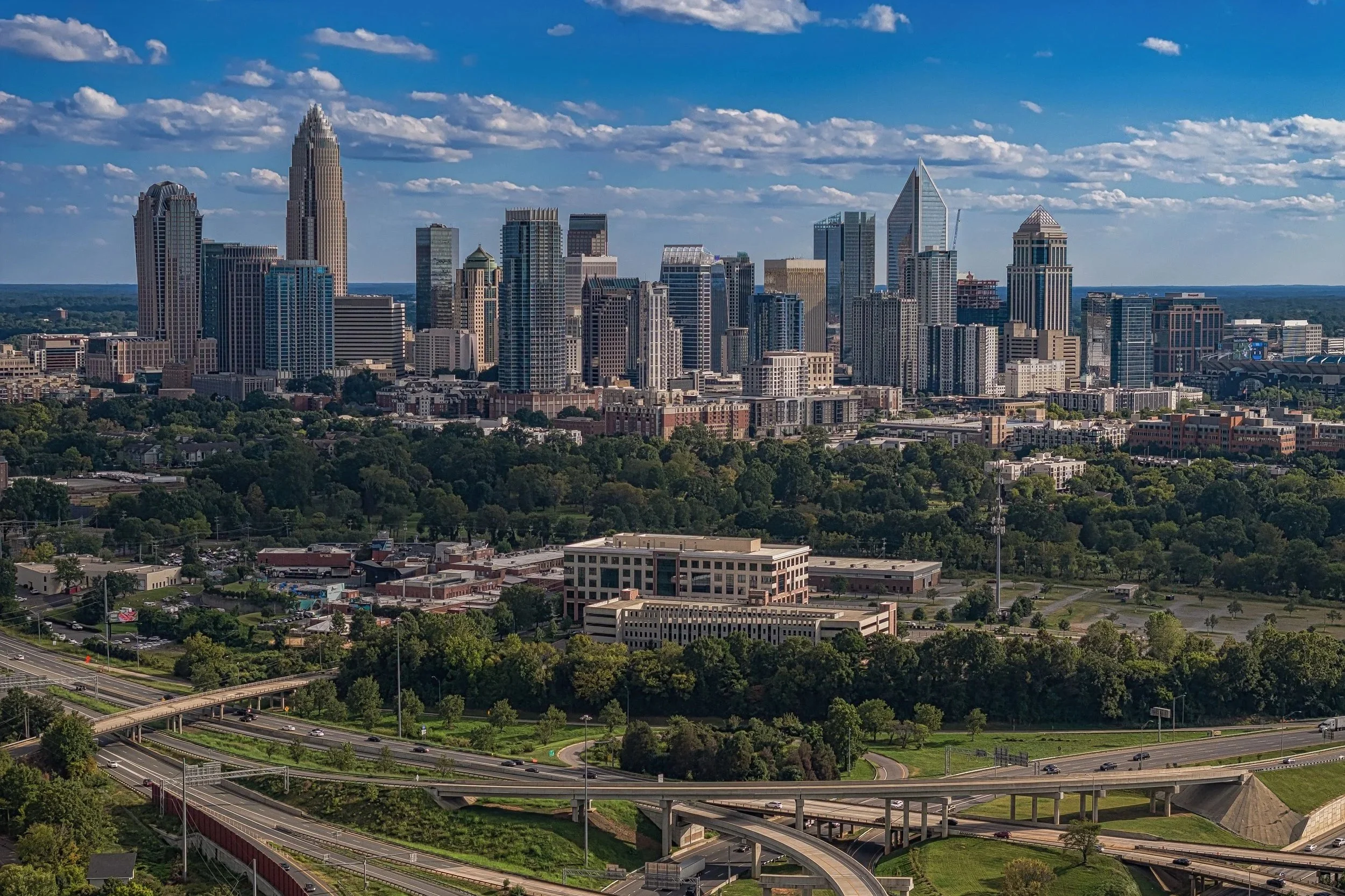 A city skyline featuring tall skyscrapers, some under construction, with a mix of modern and historic architectural styles, set against a blue sky with scattered clouds. The foreground includes green trees, roads, and highway overpasses.