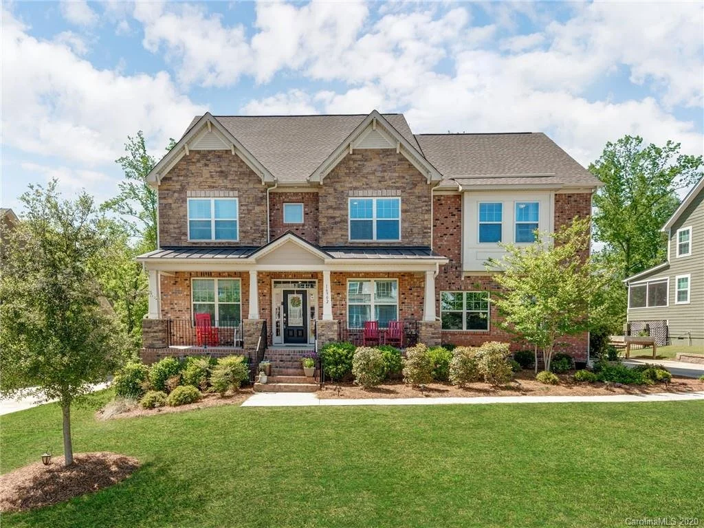 A two-story brick house with a front porch, two red chairs, and mature trees in the yard under a partly cloudy sky.