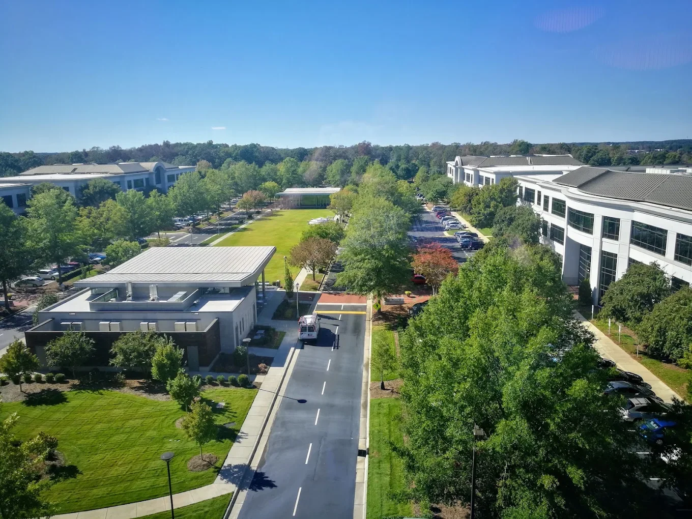 Bird's eye view of a modern campus with white office buildings, parking lots, landscaped green spaces, and trees on a sunny day.