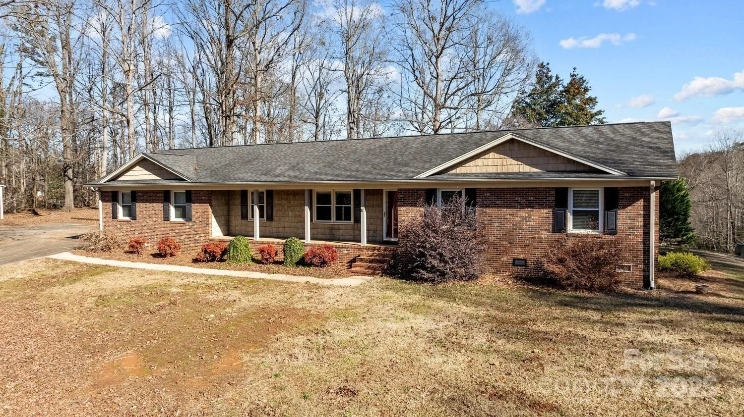 Single-story brick house with front porch, surrounded by lawn, shrubs, and trees in the background, under a blue sky with clouds.