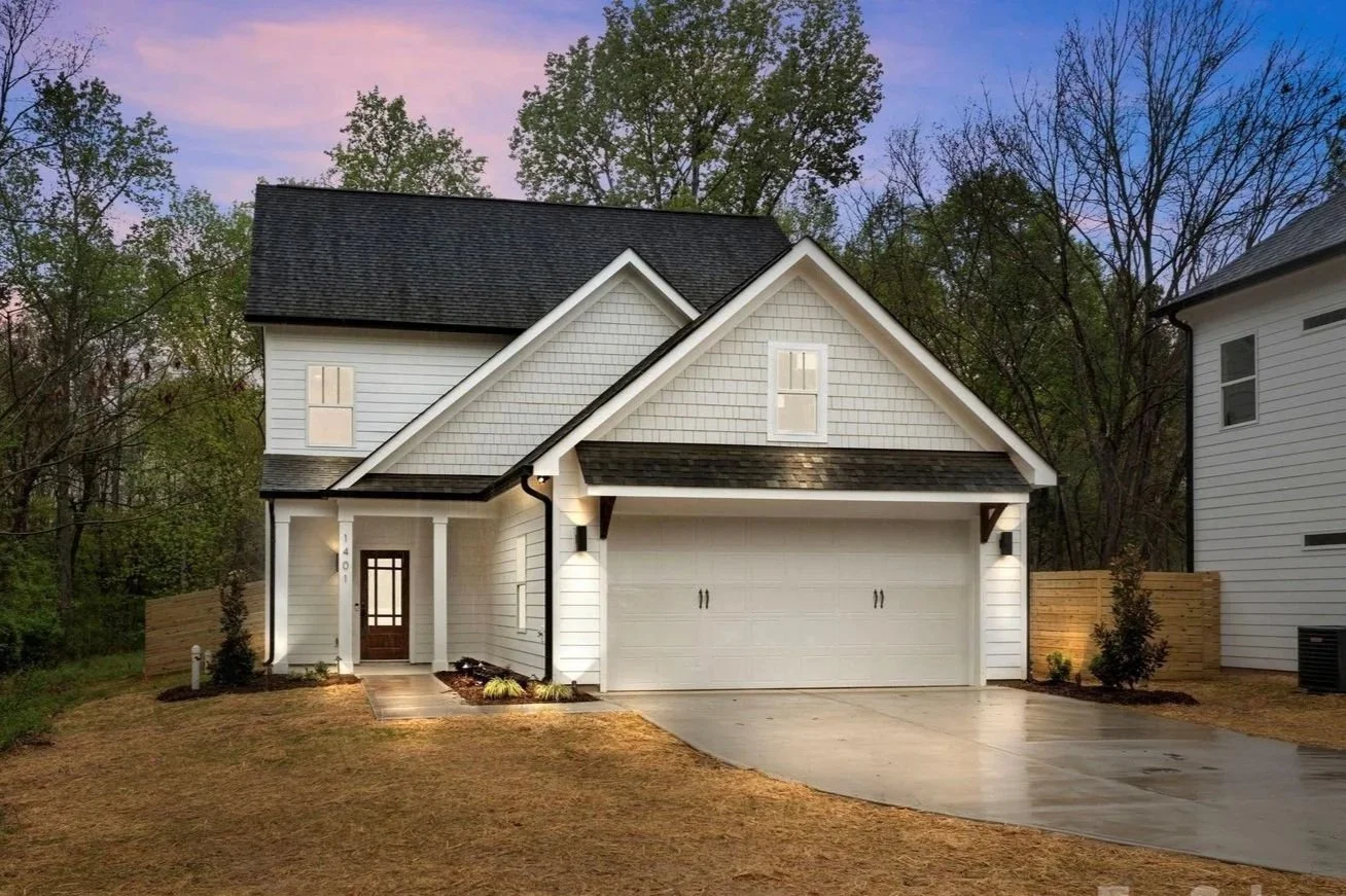 A modern two-story white house with a black roof, attached garage, small porch with a wooden door, surrounded by a lawn and a wooden fence, during dusk.