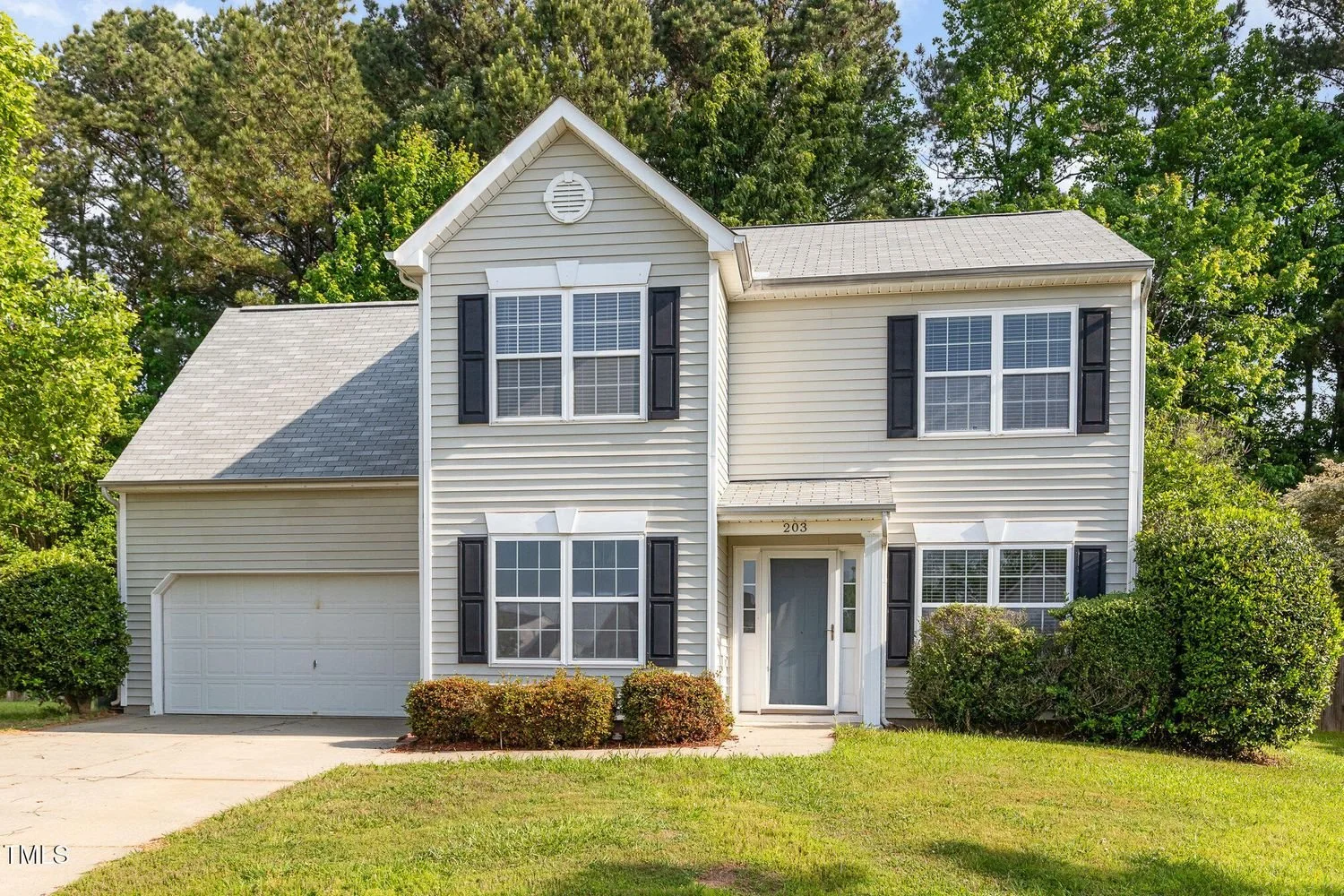 A two-story suburban house with beige siding, black shutters, and a gray roof, surrounded by green bushes and trees.