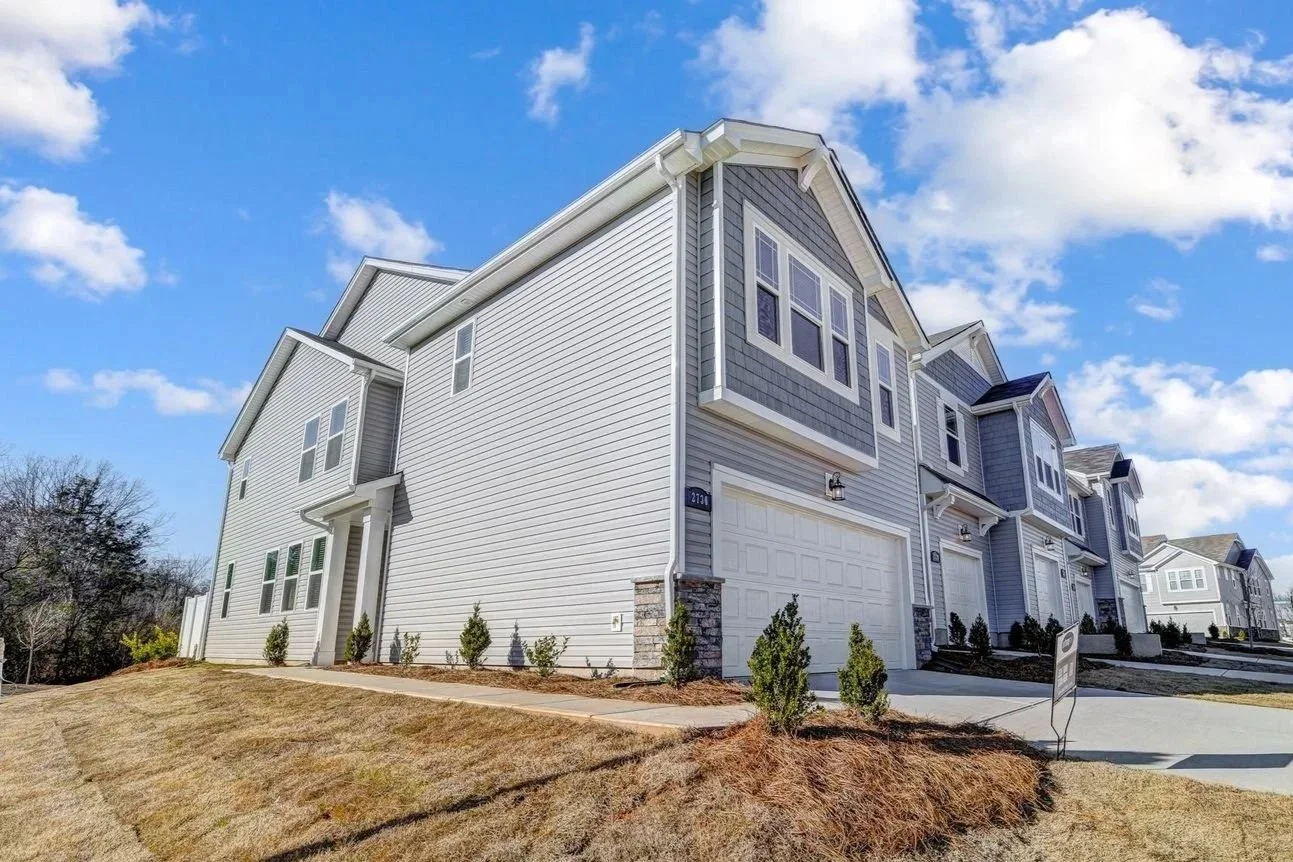 A row of new townhouses with gray and white siding, garages, and small lawns under a blue sky with scattered clouds.