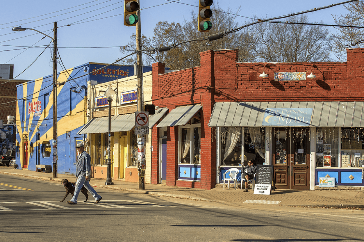 Street view showing buildings with colorful storefronts, a man walking a dog across crosswalk, traffic lights, and utility poles.
