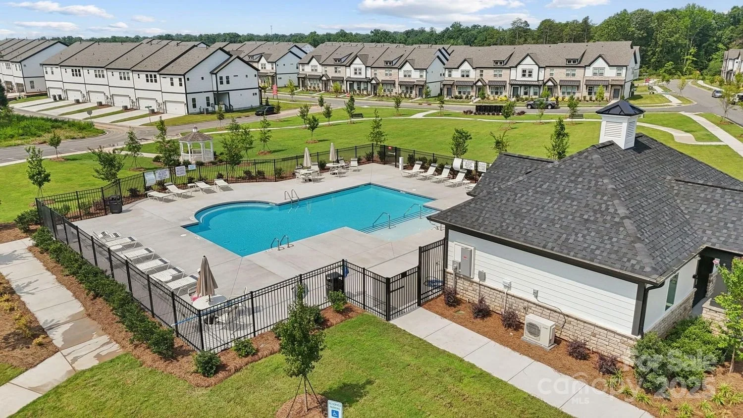 A community swimming pool with lounge chairs and umbrellas, surrounded by a black fence, with a small building nearby. In the background, there are residential houses and green trees.