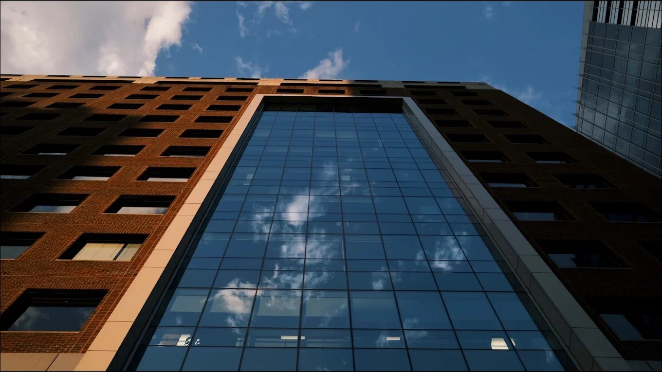 Low-angle view of a tall building with a glass facade that reflects the sky and clouds, flanked by brick and glass structures.