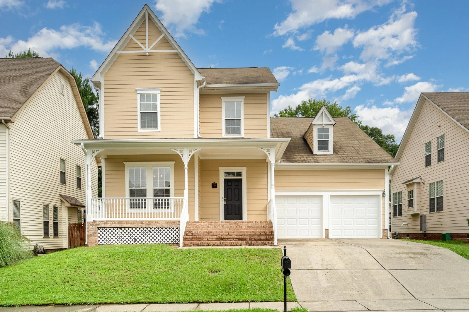 Cute two-story yellow house with white trim and a black front door, brick steps, a porch with white railing, attached double garage, green lawn, blue sky with white clouds.