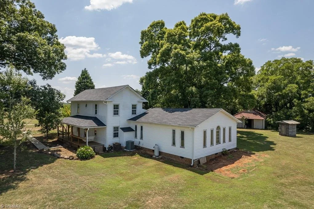 A white two-story house with a wrap-around porch, surrounded by a grassy yard and trees, under a partly cloudy sky.