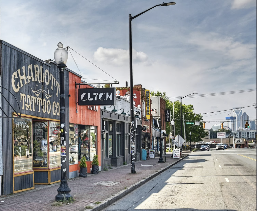 Empty city street with storefronts and signs, including Charlotte Tattoo Co and Clutch, with cars at a traffic light and tall buildings in the distance under a partly cloudy sky.