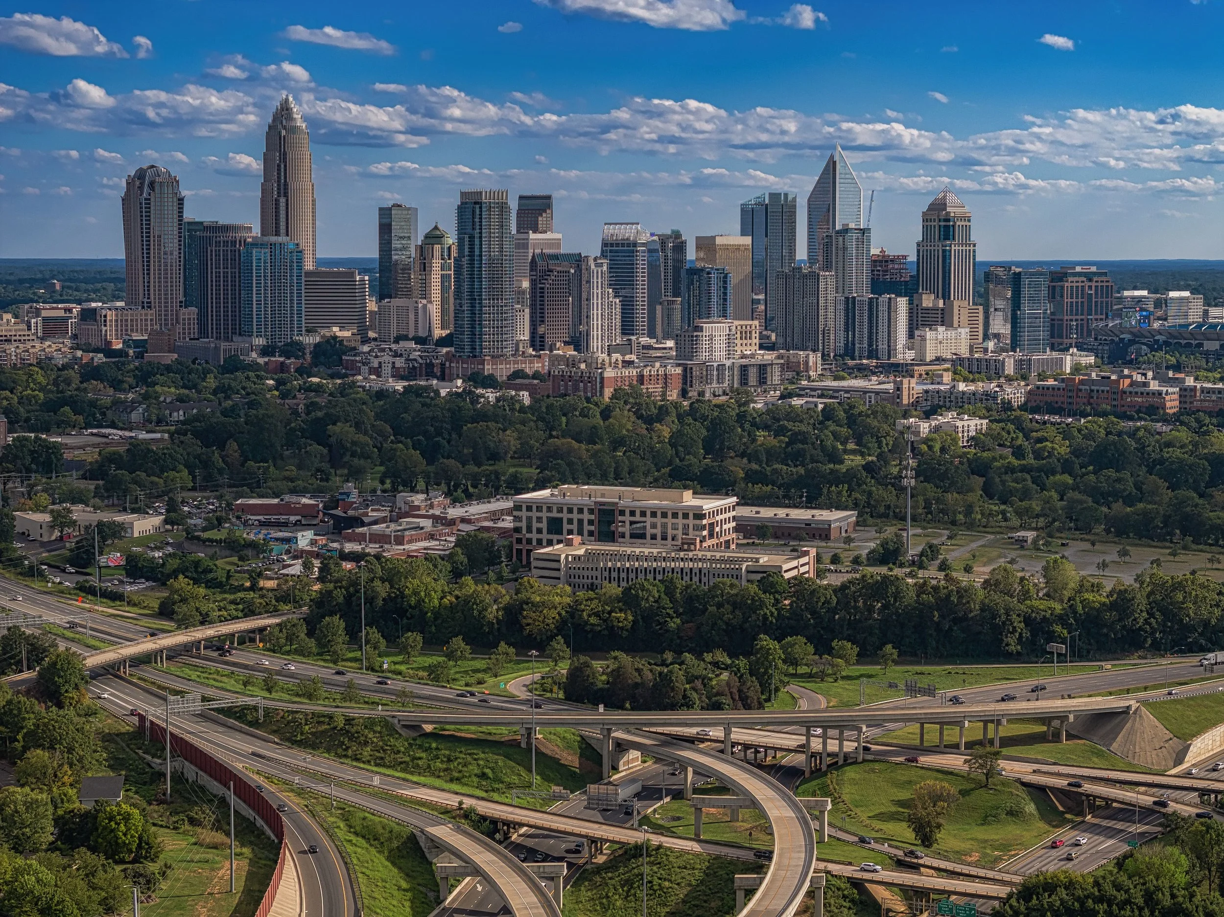 A city skyline with tall skyscrapers under a partly cloudy sky, with highways and green areas in the foreground.