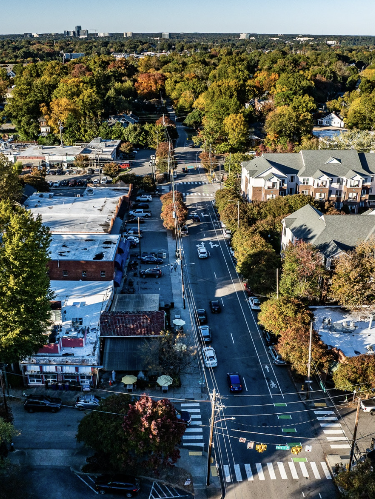 An aerial view of a city street lined with cars, trees with colorful autumn foliage, and buildings including a commercial area and residential apartments, with a park visible in the distance.