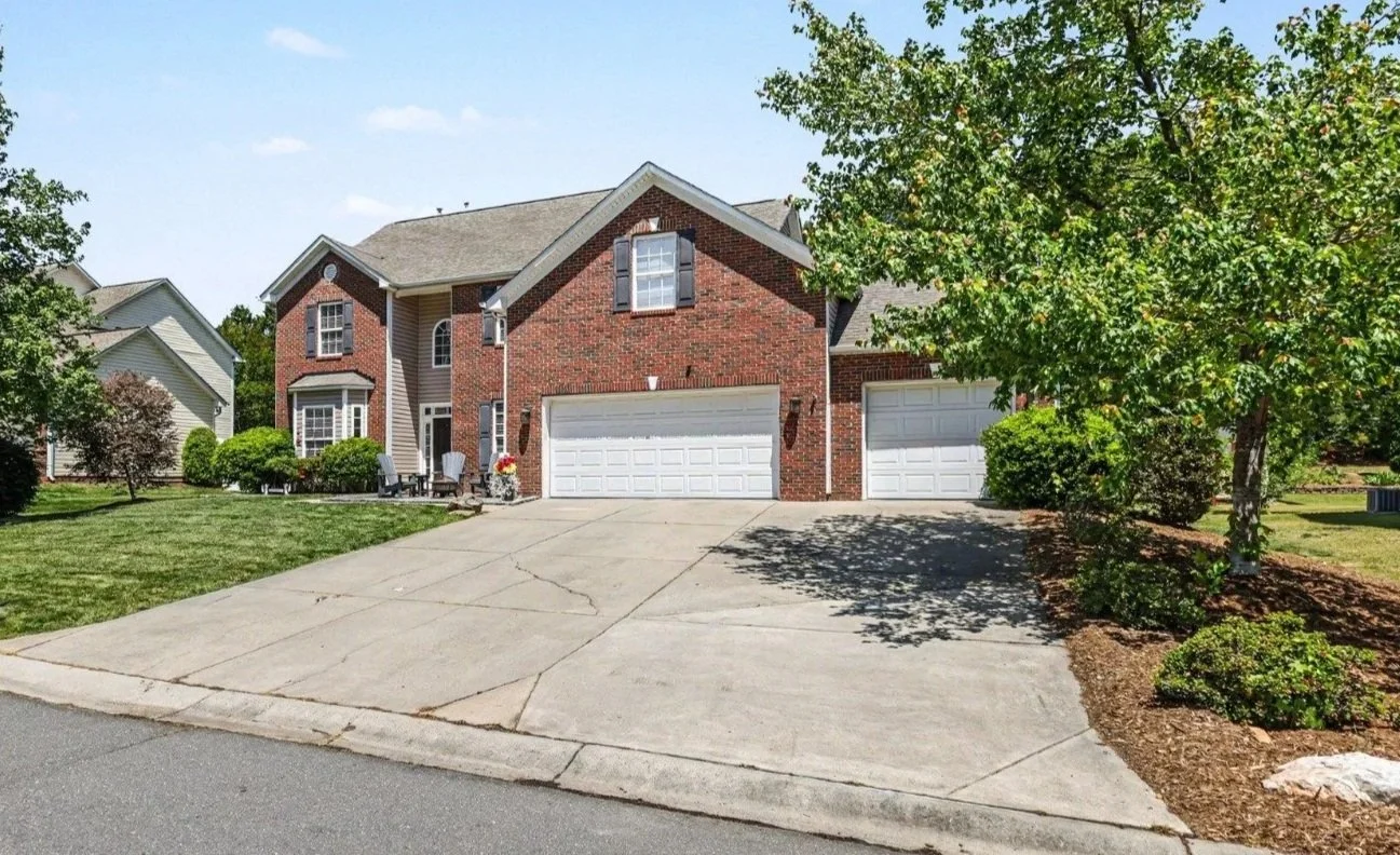 A two-story brick house with attached two-car garage, front porch, bay window, black shutters, and landscaped front yard.