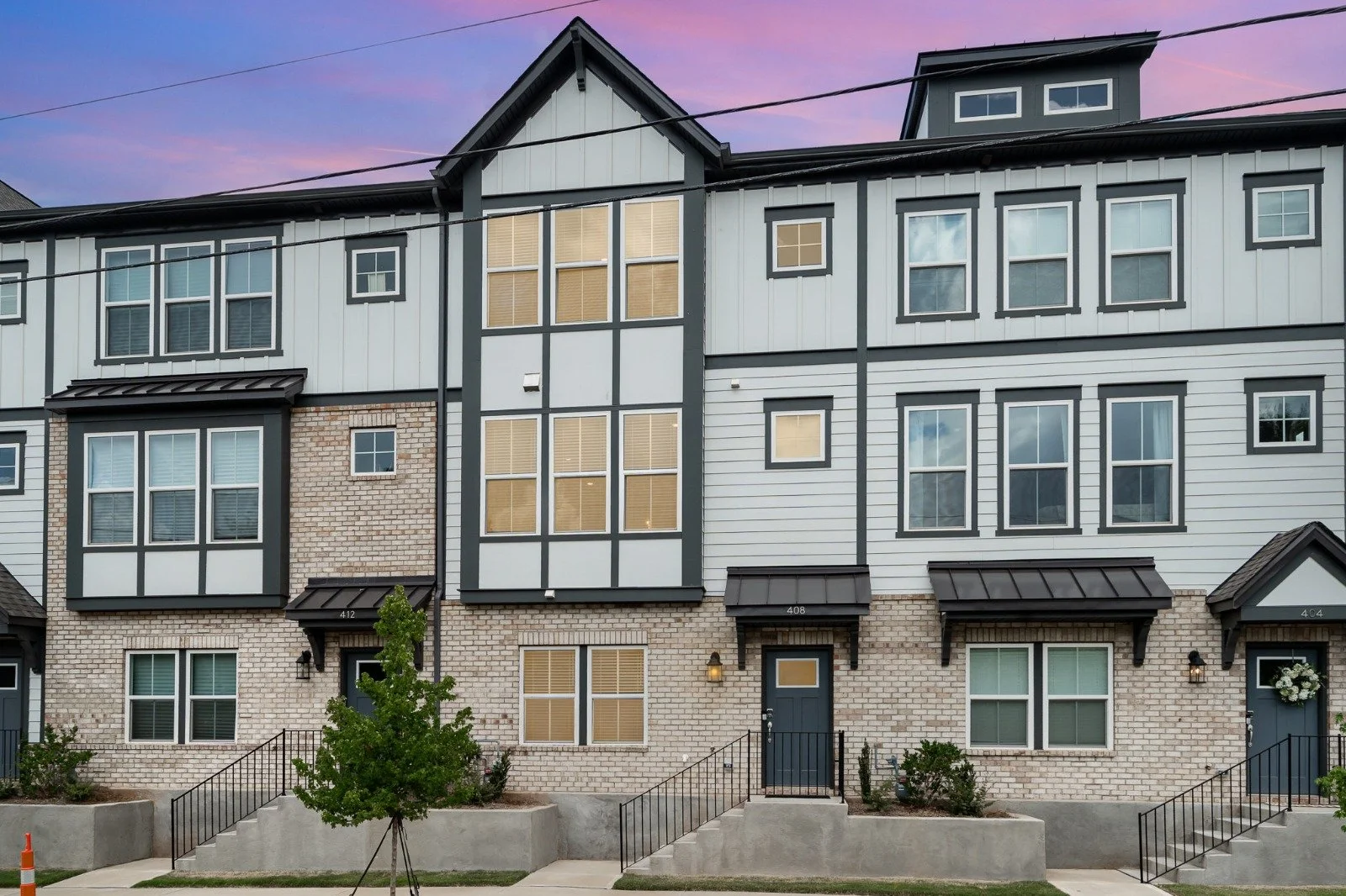 Multi-story modern townhouses with brick and siding exteriors, several windows, cornices, and front steps, under a colorful sunset sky.