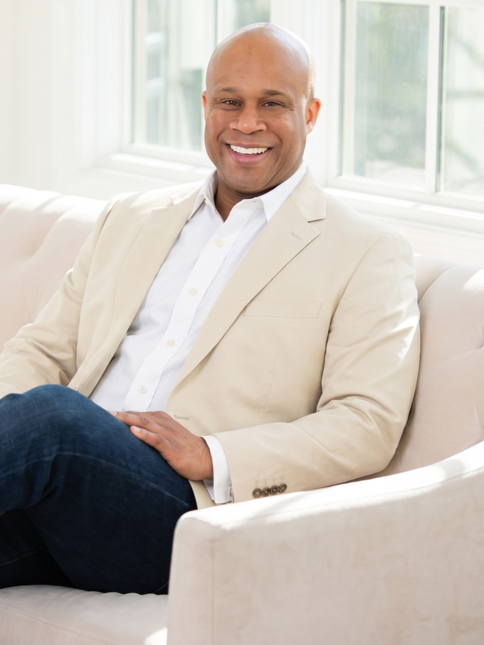 A smiling man with a shaved head sitting on a cream-colored sofa near large windows, wearing a white shirt and beige blazer.