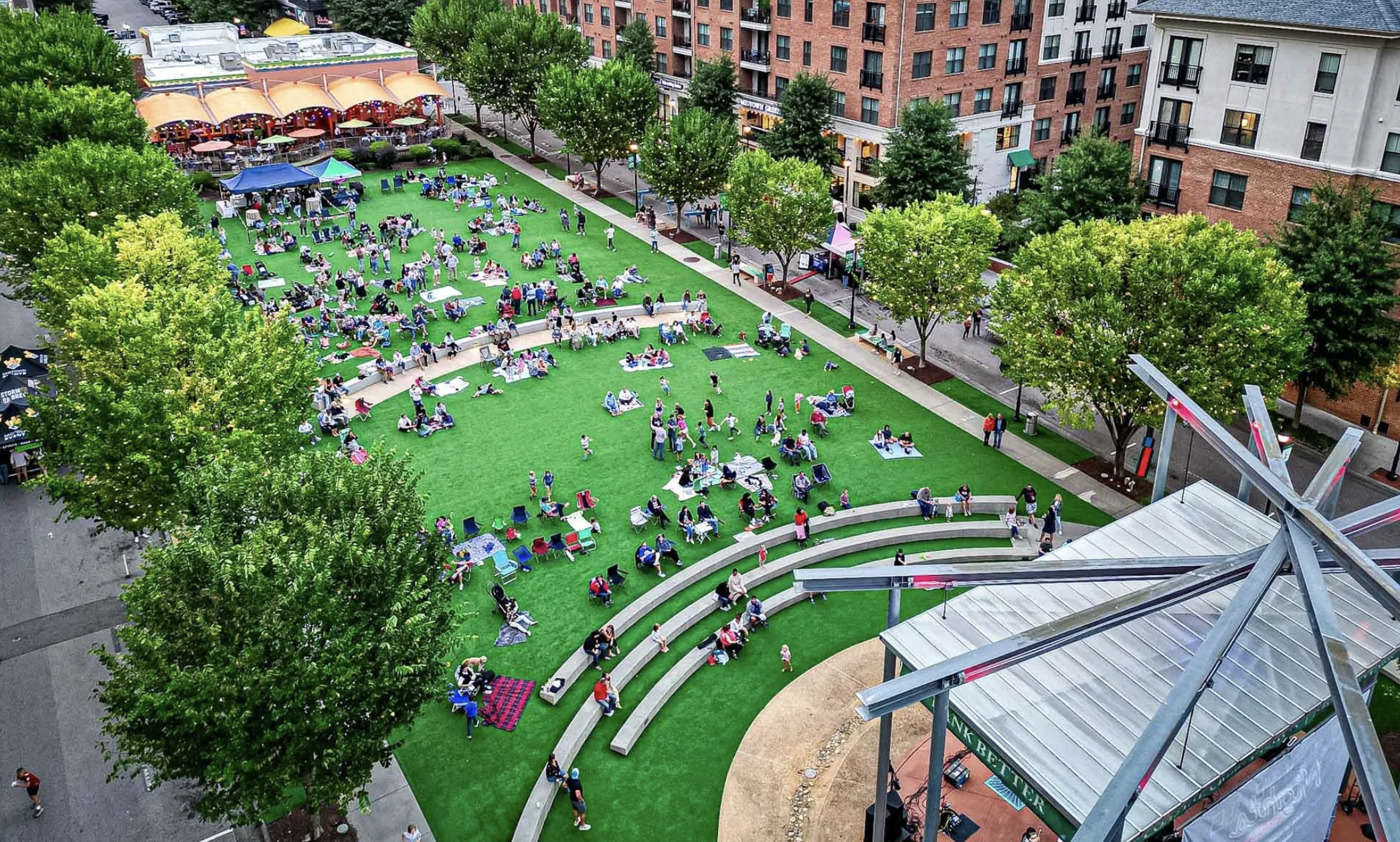 Bird's eye view of a lively urban park with many people relaxing on blankets and sitting on benches, surrounded by trees, a curved stage, and nearby buildings, with street and sidewalk areas.