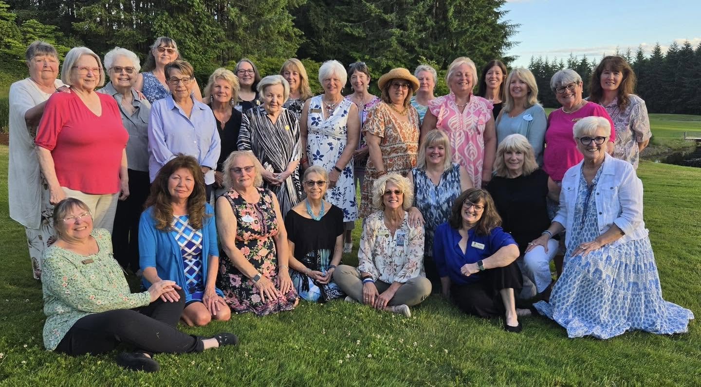 A group of around 30 women outdoors on a grassy field with trees in the background during daytime.