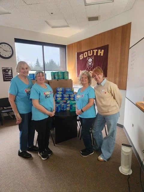 Four women standing behind a table with packages of diapers, in a room with a South school banner on the wall.
